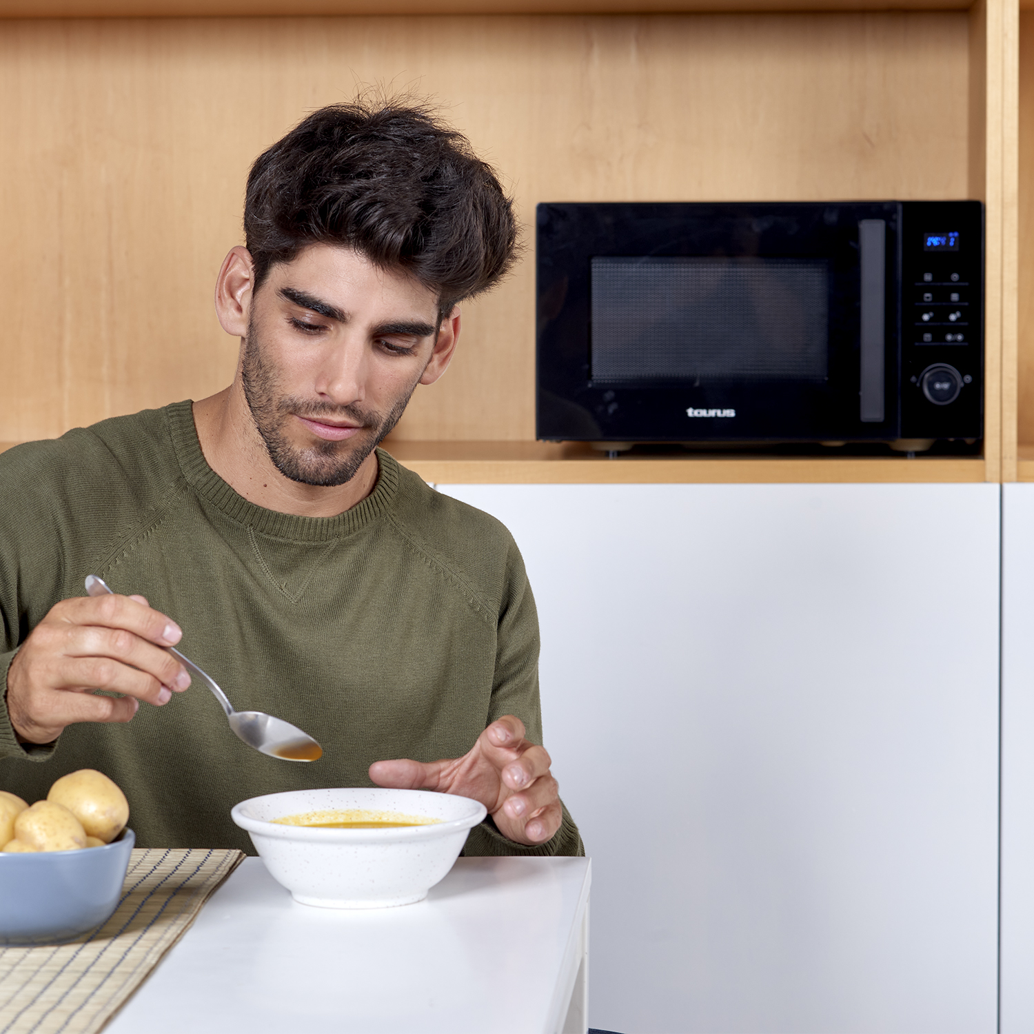 Hombre comiendo sopa, tazón de sopa en primer plano, microondas al fondo.
