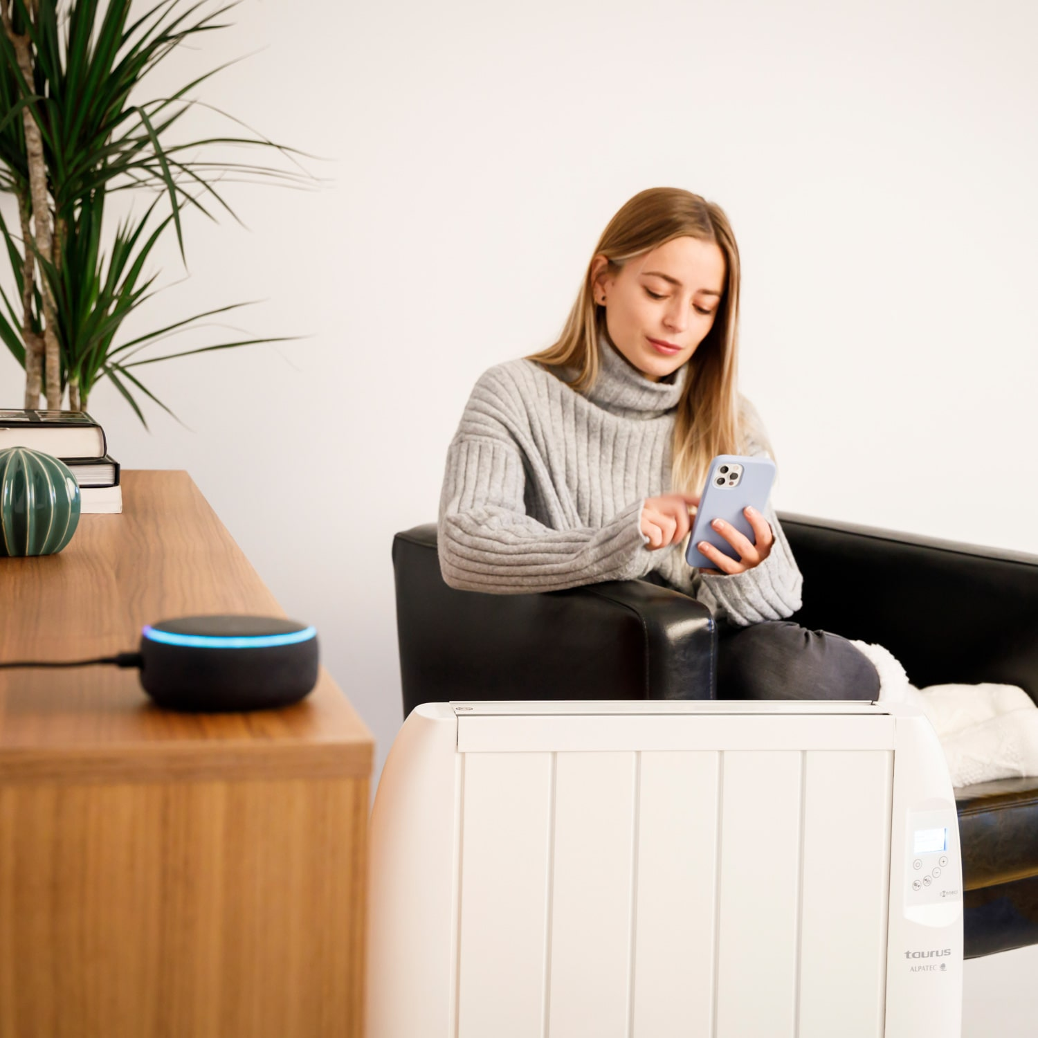 Una mujer usa un teléfono inteligente mientras está sentada en un sofá negro. Un altavoz Alexa está en la mesa auxiliar.