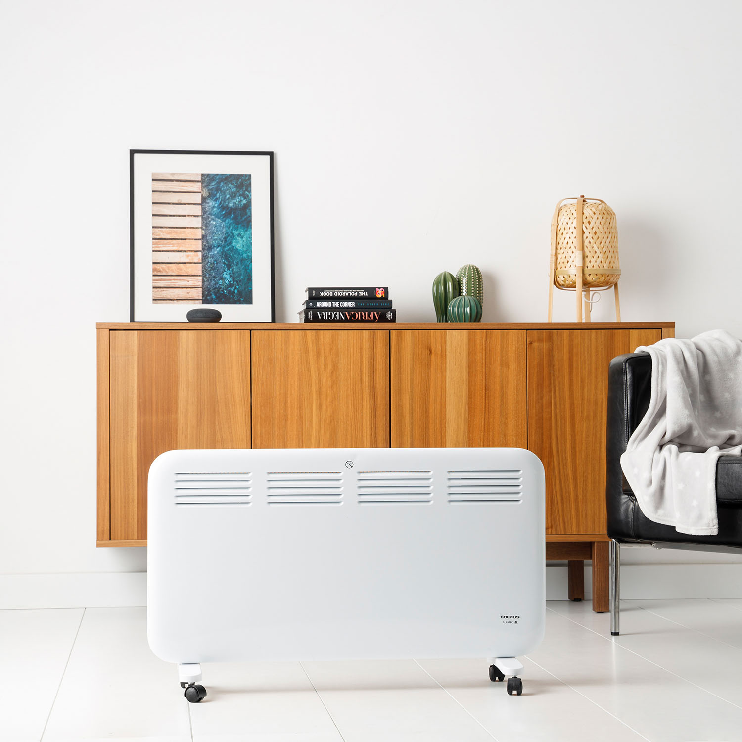 Un calefactor eléctrico blanco sobre ruedas está frente a un armario con una imagen y lámpara al lado.