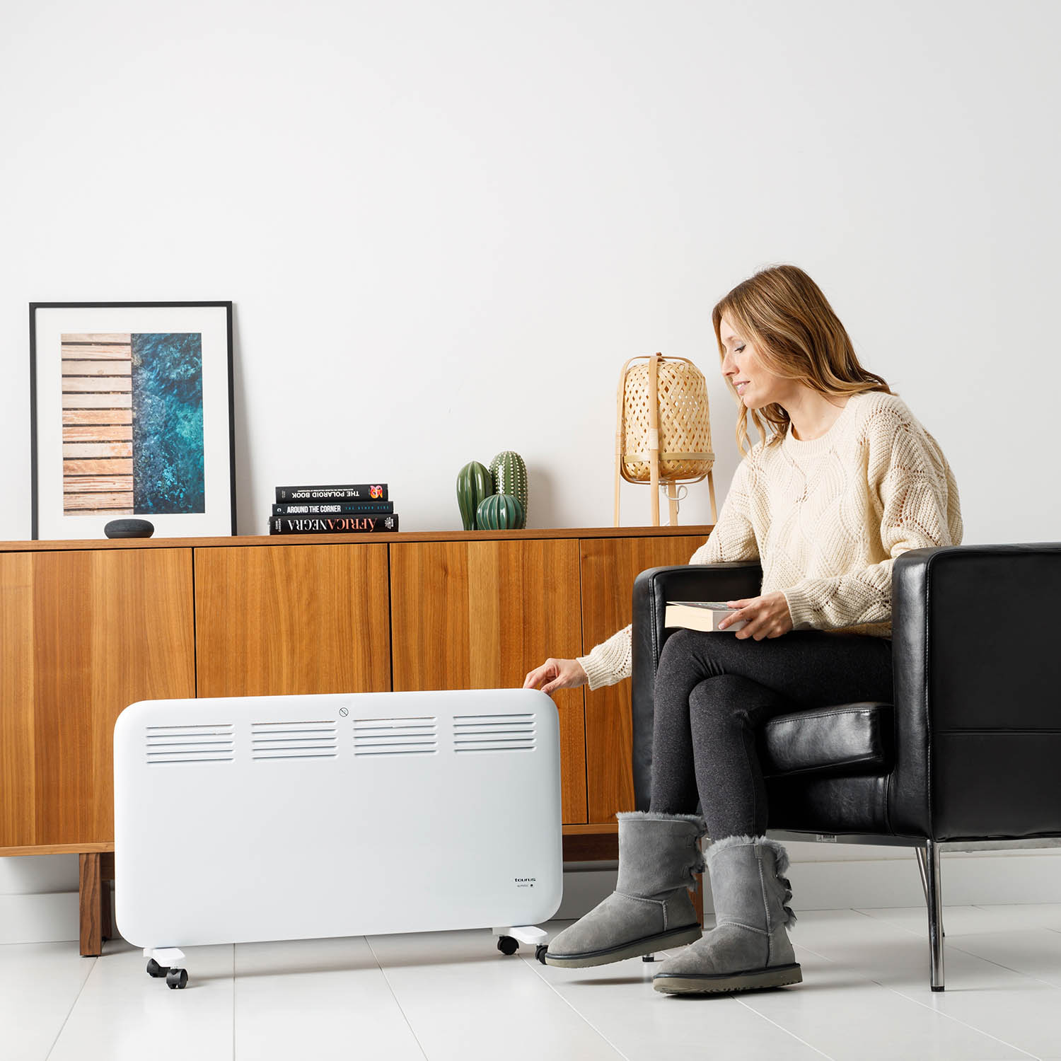 Una mujer sentada en una habitación con un calefactor blanco sobre ruedas, leyendo un libro y cerca de un mueble de madera.