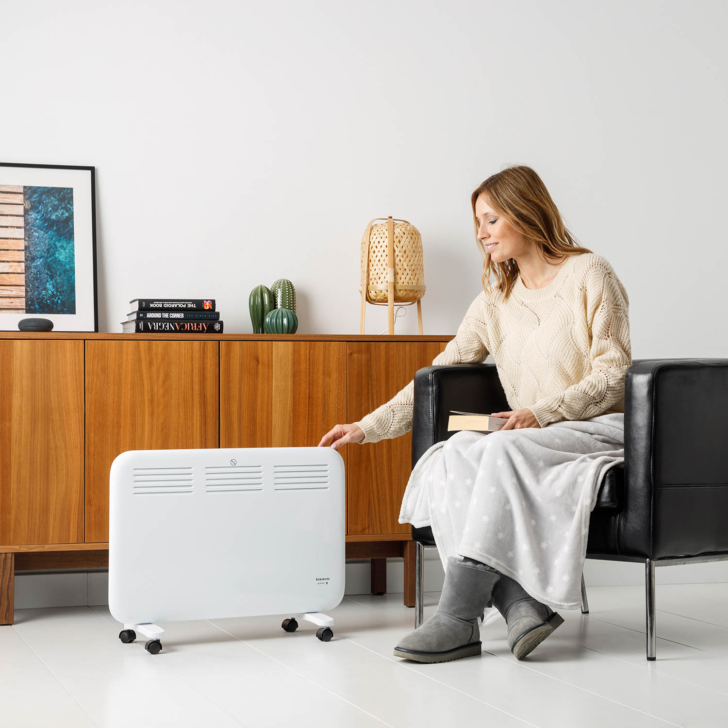 Mujer con libro junto a calefactor blanco con ruedas. Gabinete de madera a la izquierda.