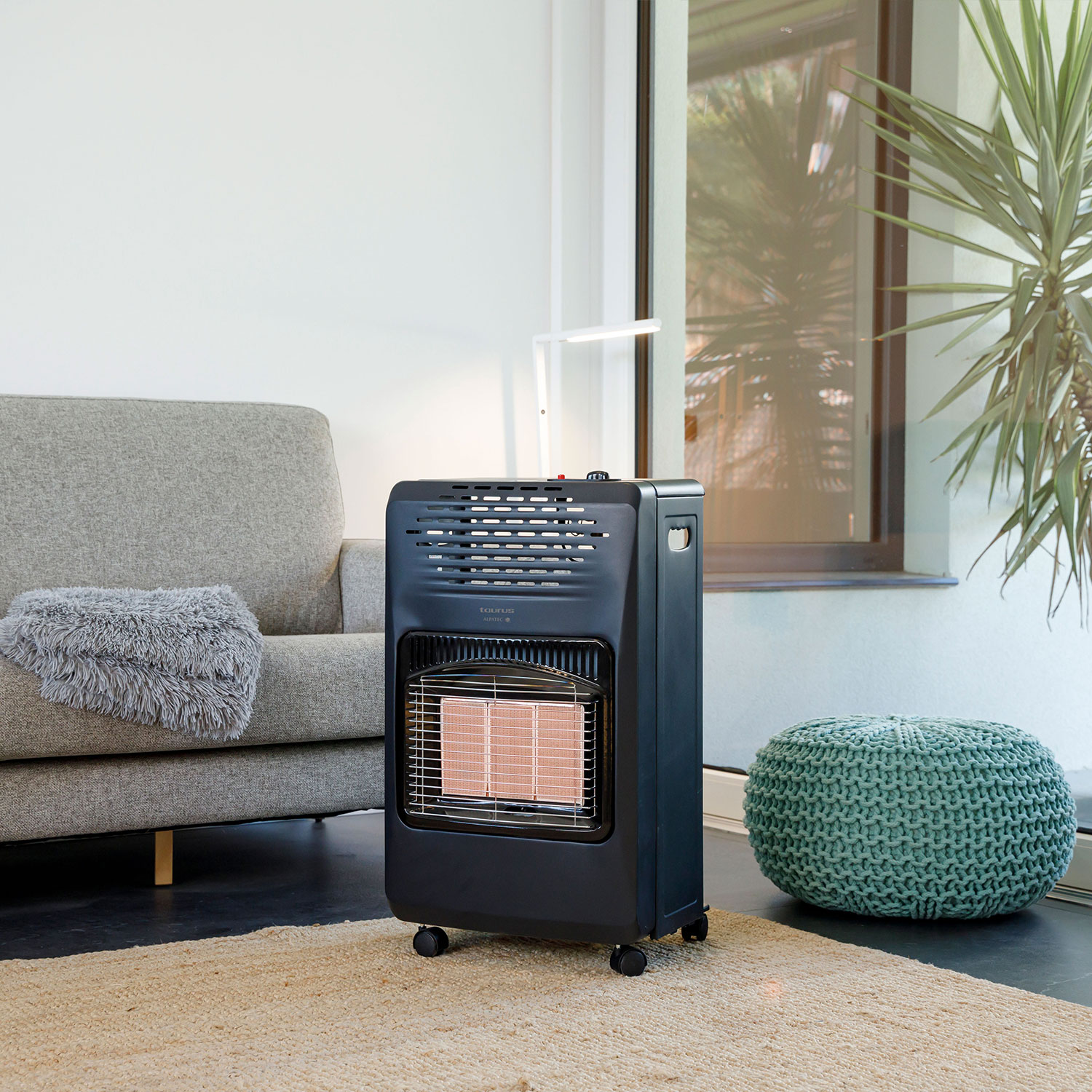 Un calefactor negro con ruedas está en una habitación. Un sofá gris y un puf verde también son visibles.
