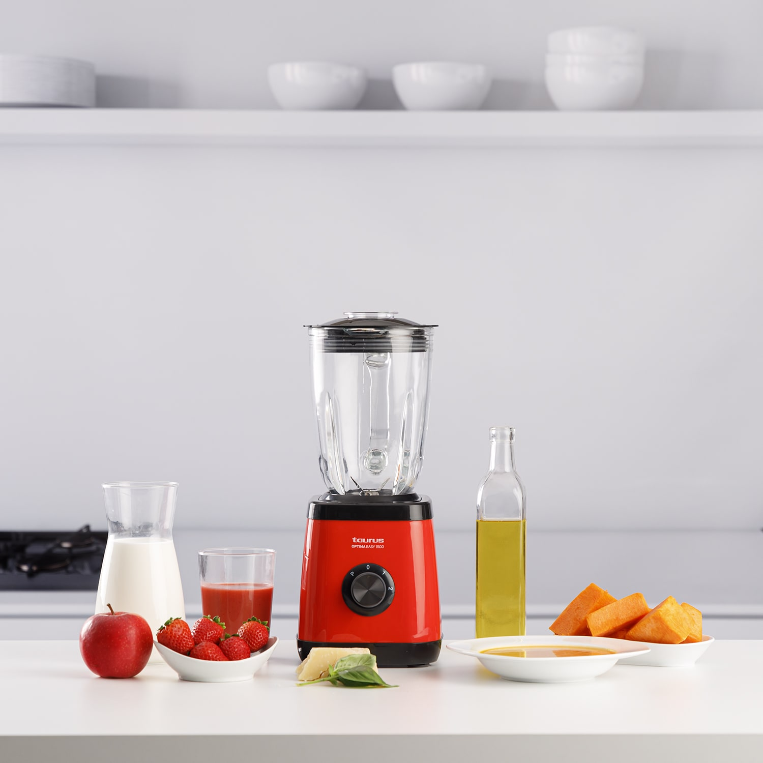 Una licuadora roja con ingredientes como leche, fresas, manzanas, aceite y calabaza sobre una superficie blanca.