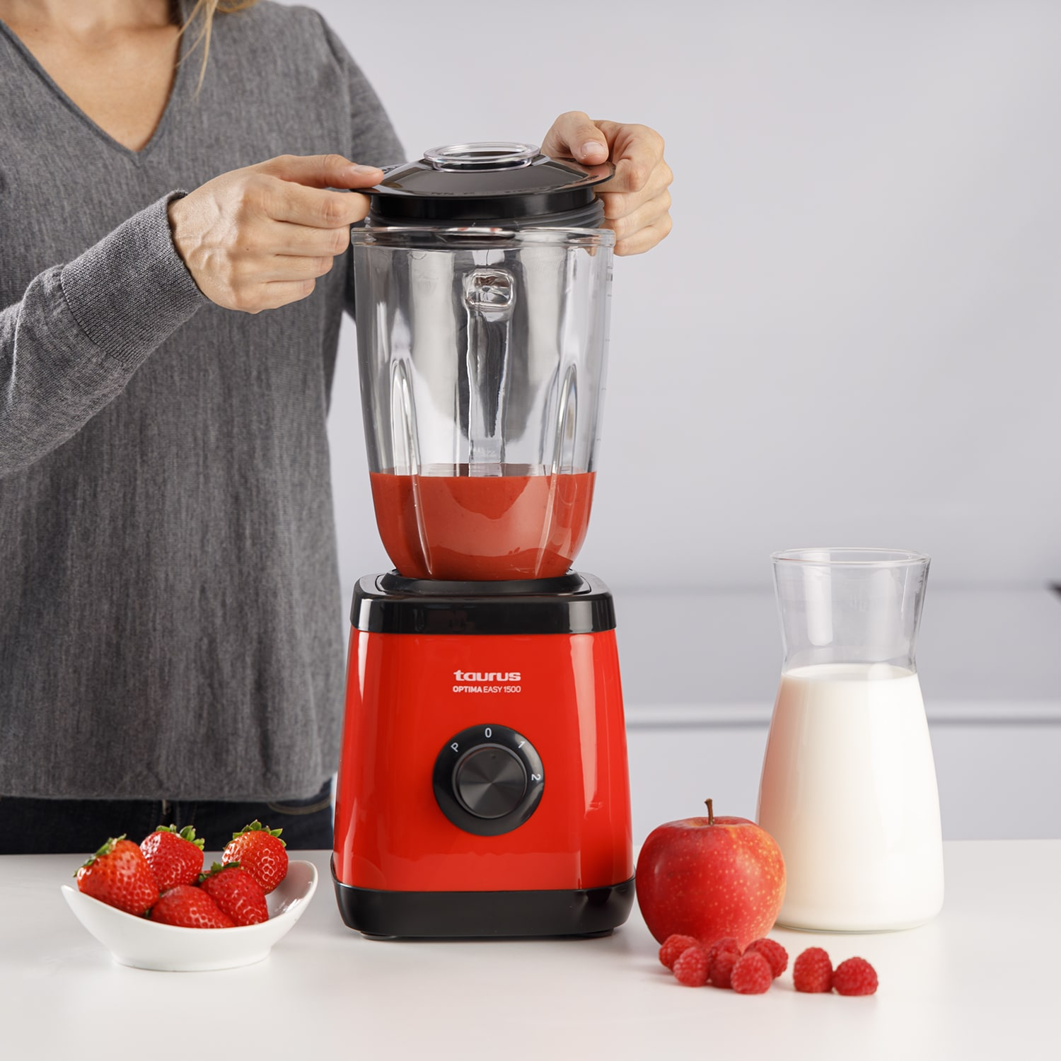 Una persona abriendo una licuadora, con fresas, frambuesas y una manzana en el mostrador.