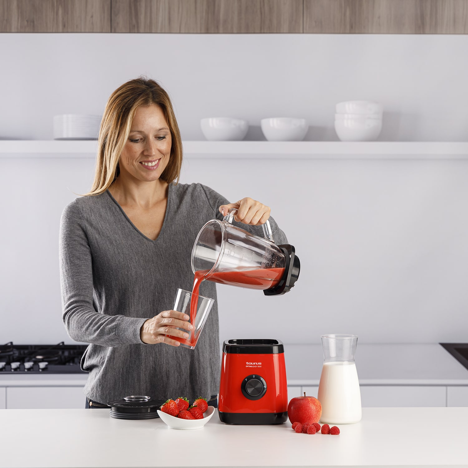 Mujer vertiendo líquido rojo de una licuadora en un vaso, se ven fresas y manzana.
