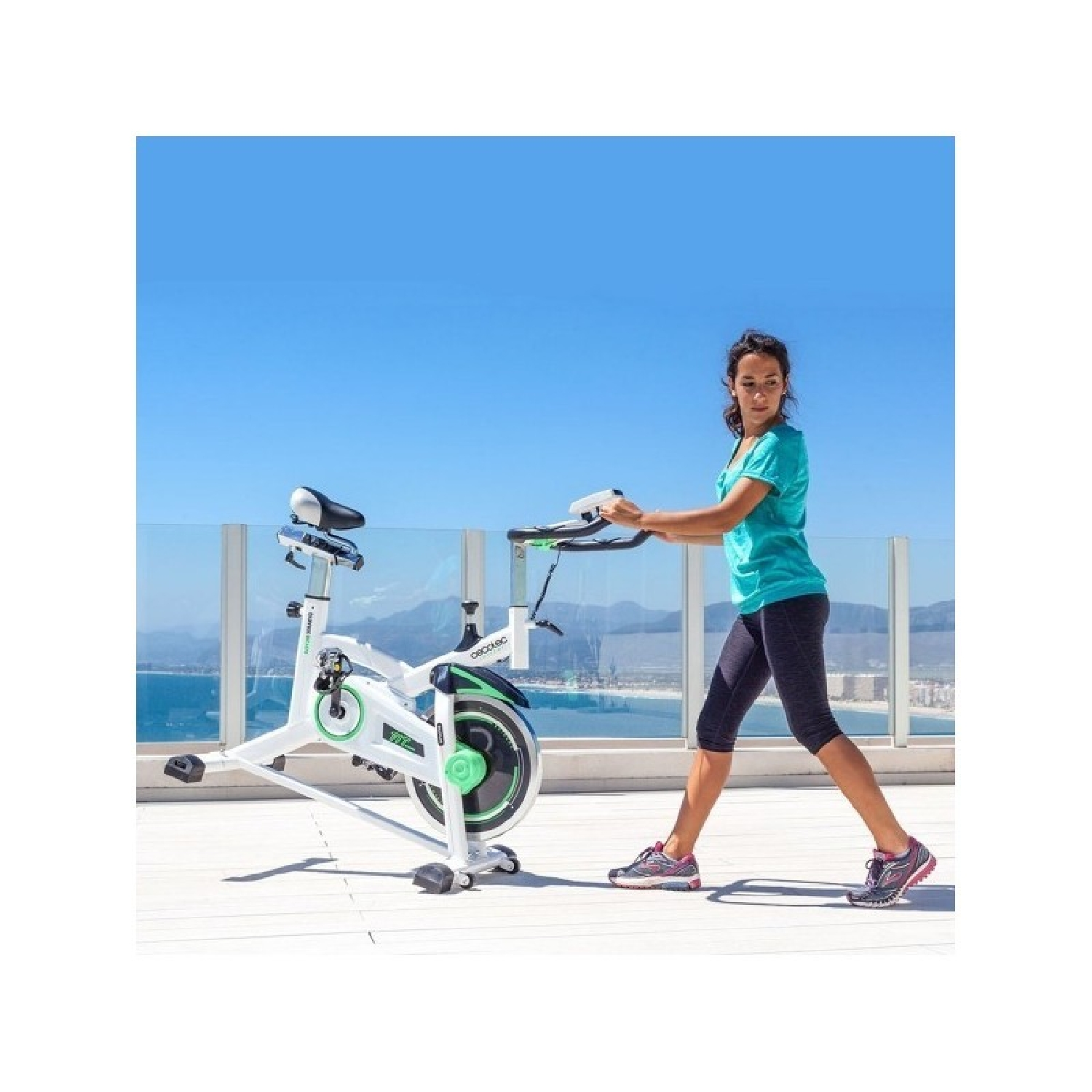 Una mujer en una bicicleta estática blanca, al aire libre con cielo azul y océano visible.