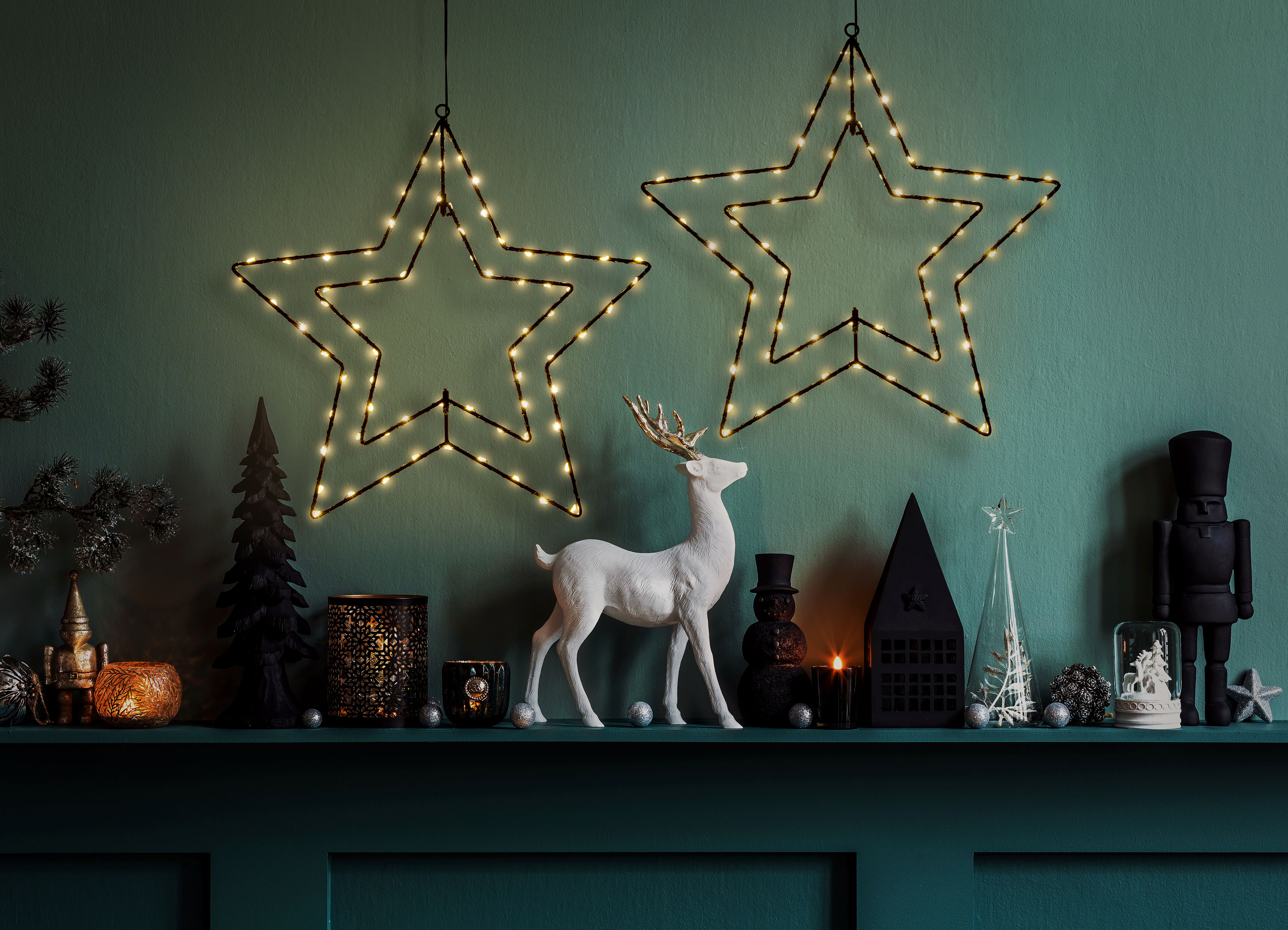 Weihnachtsdekorationen an einer grünen Wand, darunter Sterne, ein Reh und Kerzen.
