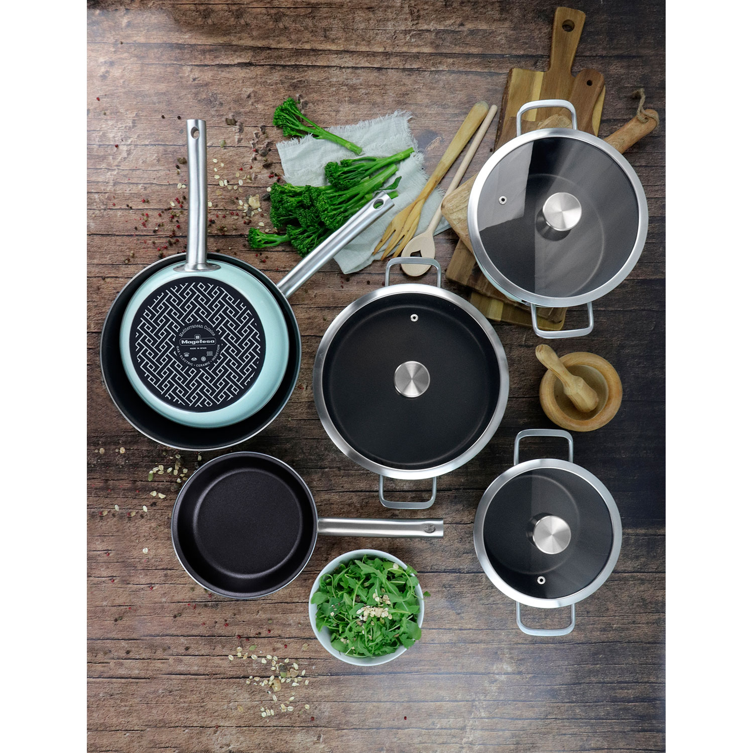 Ein Set verschiedener Pfannen und Töpfe auf Holz, mit Gemüse und Küchenutensilien.