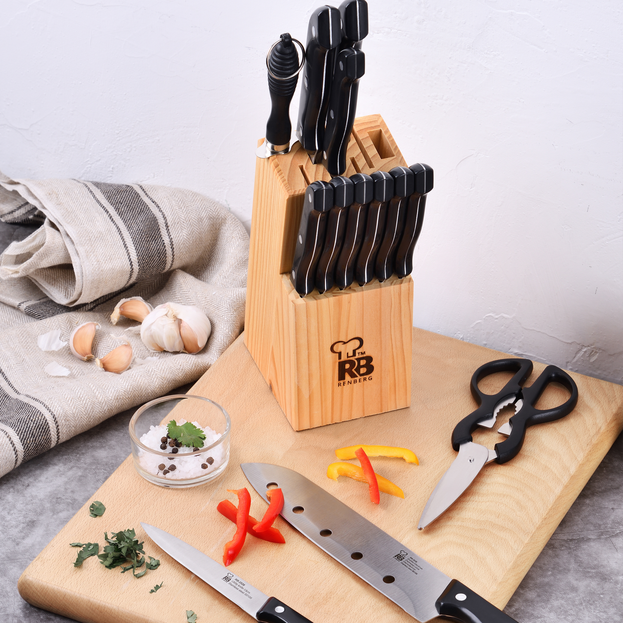 Cuchillos en bloque de madera, tabla de cortar con ingredientes, especias y tijeras, todo en superficie blanca.