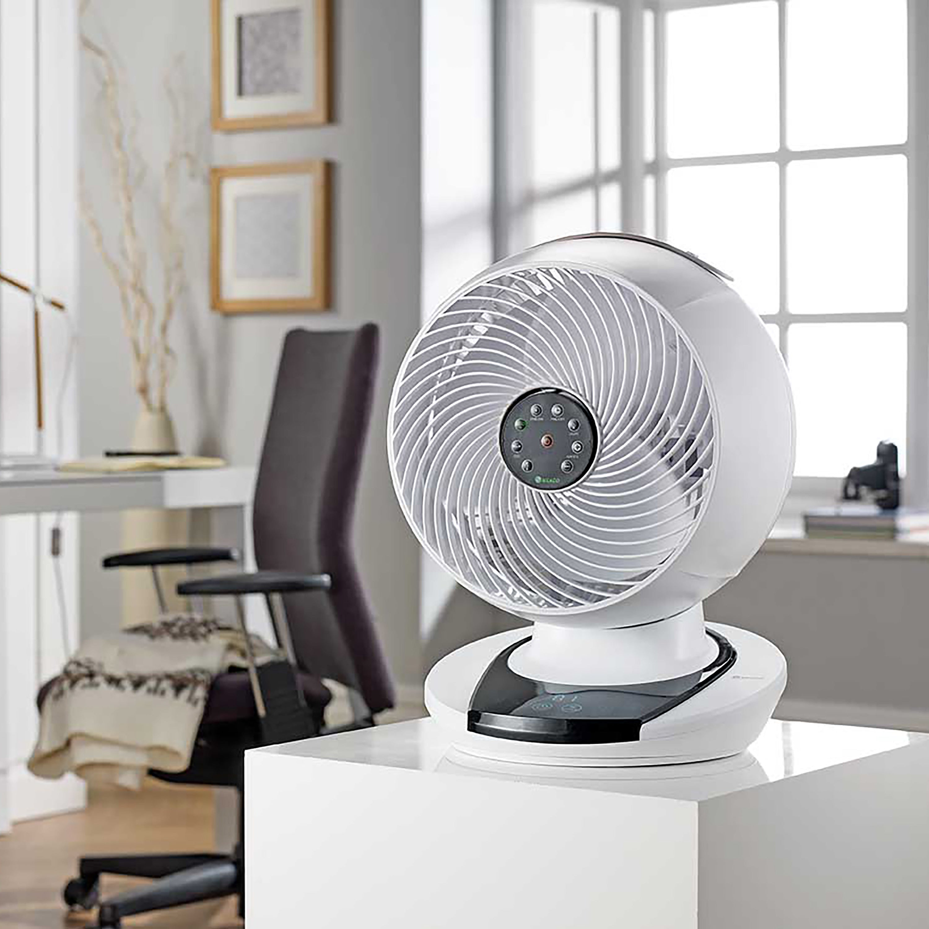 Biały wentylator na białym stojaku, z czarnym krzesłem w tle.