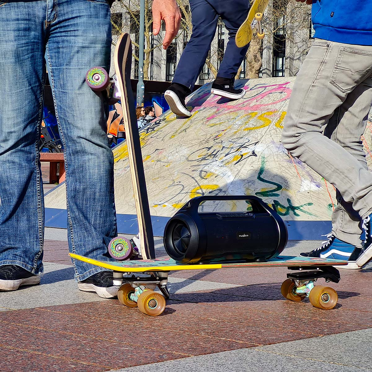 Trzy osoby i czarny głośnik na deskorolkach na rampie skateparku. Dwie osoby idą.