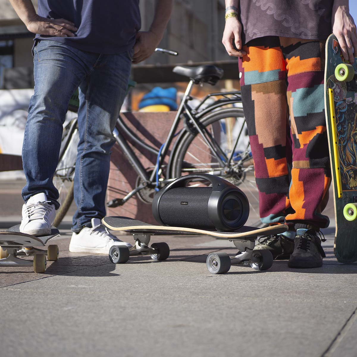 Dwie osoby ze deskorolkami i głośnikiem. Czarny głośnik stoi na deskorolce.