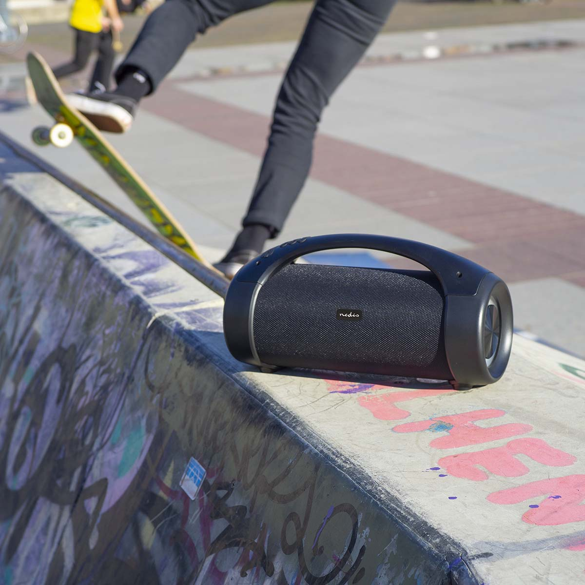 Czarny głośnik przenośny na rampie skateparku. Ktoś jeździ na deskorolce w tle.