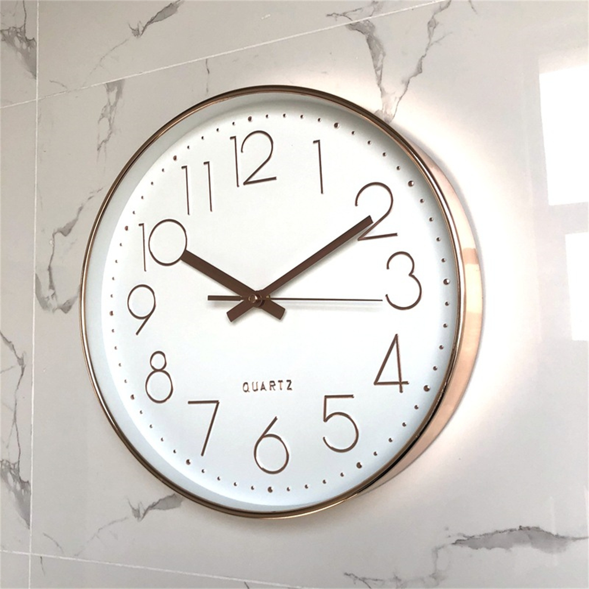 Reloj blanco con números y manecillas marrones en pared blanca tipo mármol. La hora es 2:10.