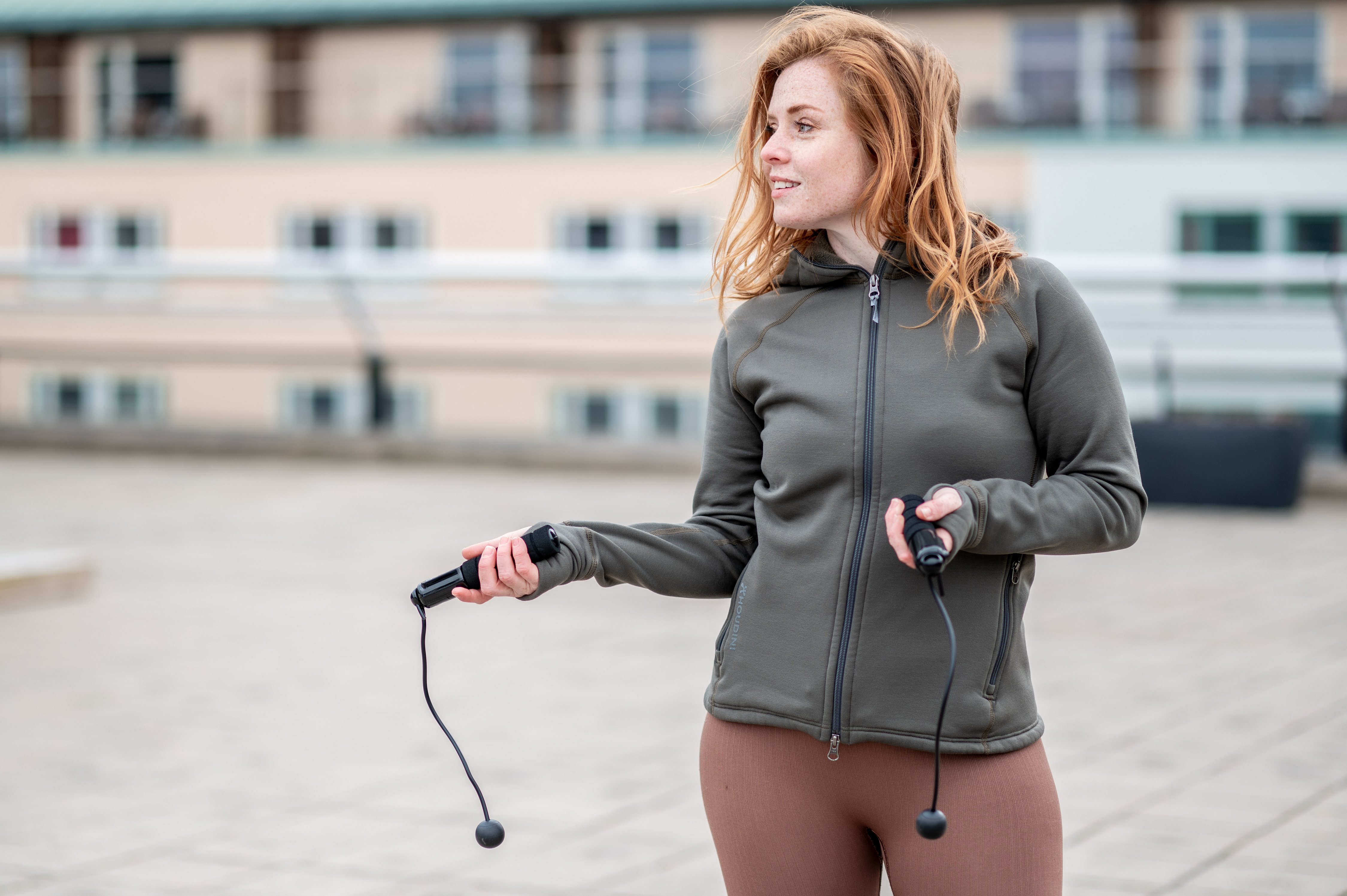 Frau in olivfarbener Jacke mit Springseil, steht auf einem Dach, blickt nach rechts.