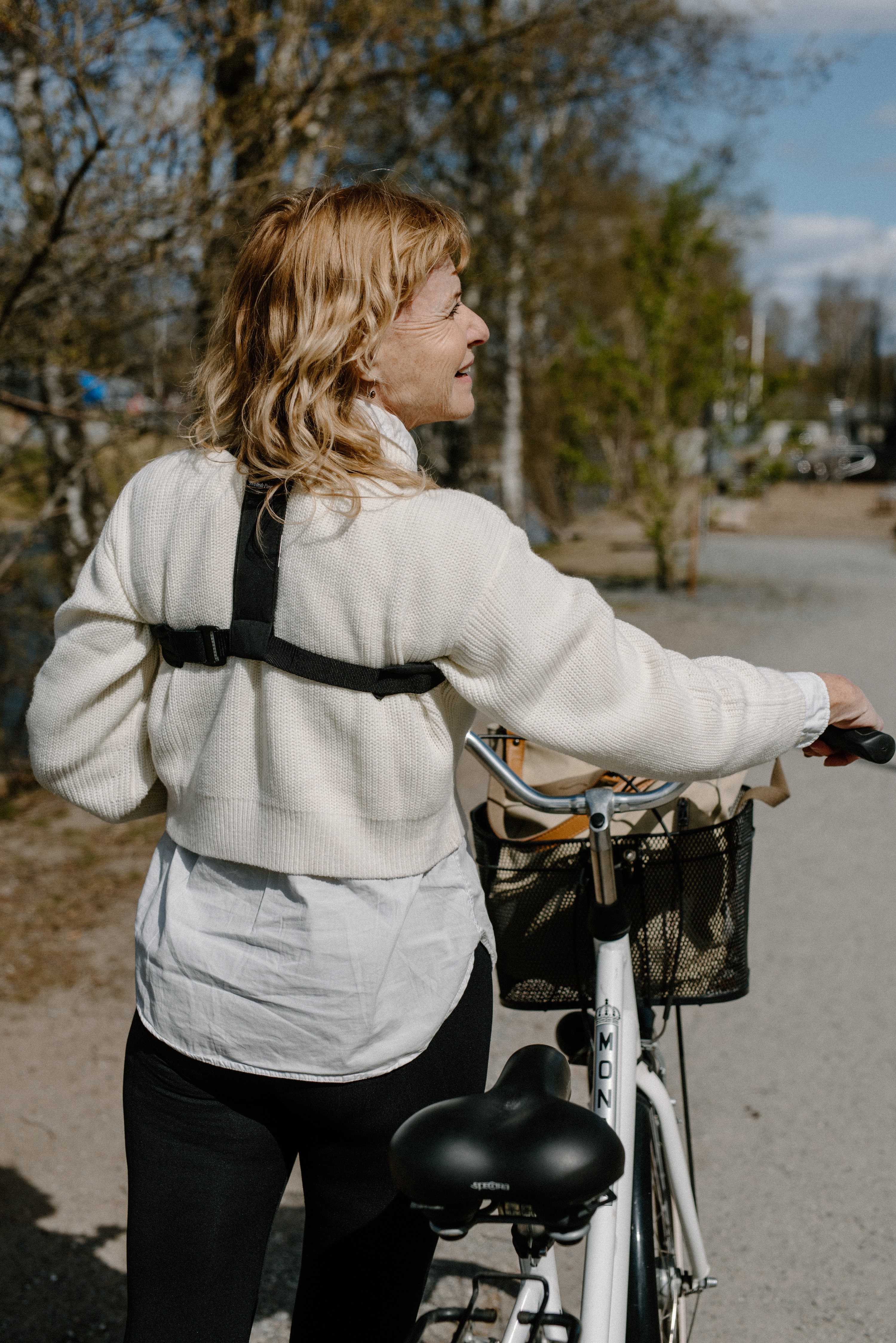 Frau mit blonden Haaren, lächelt, steht neben einem weißen Fahrrad. Sie trägt einen weißen Pullover und eine schwarze Hose.