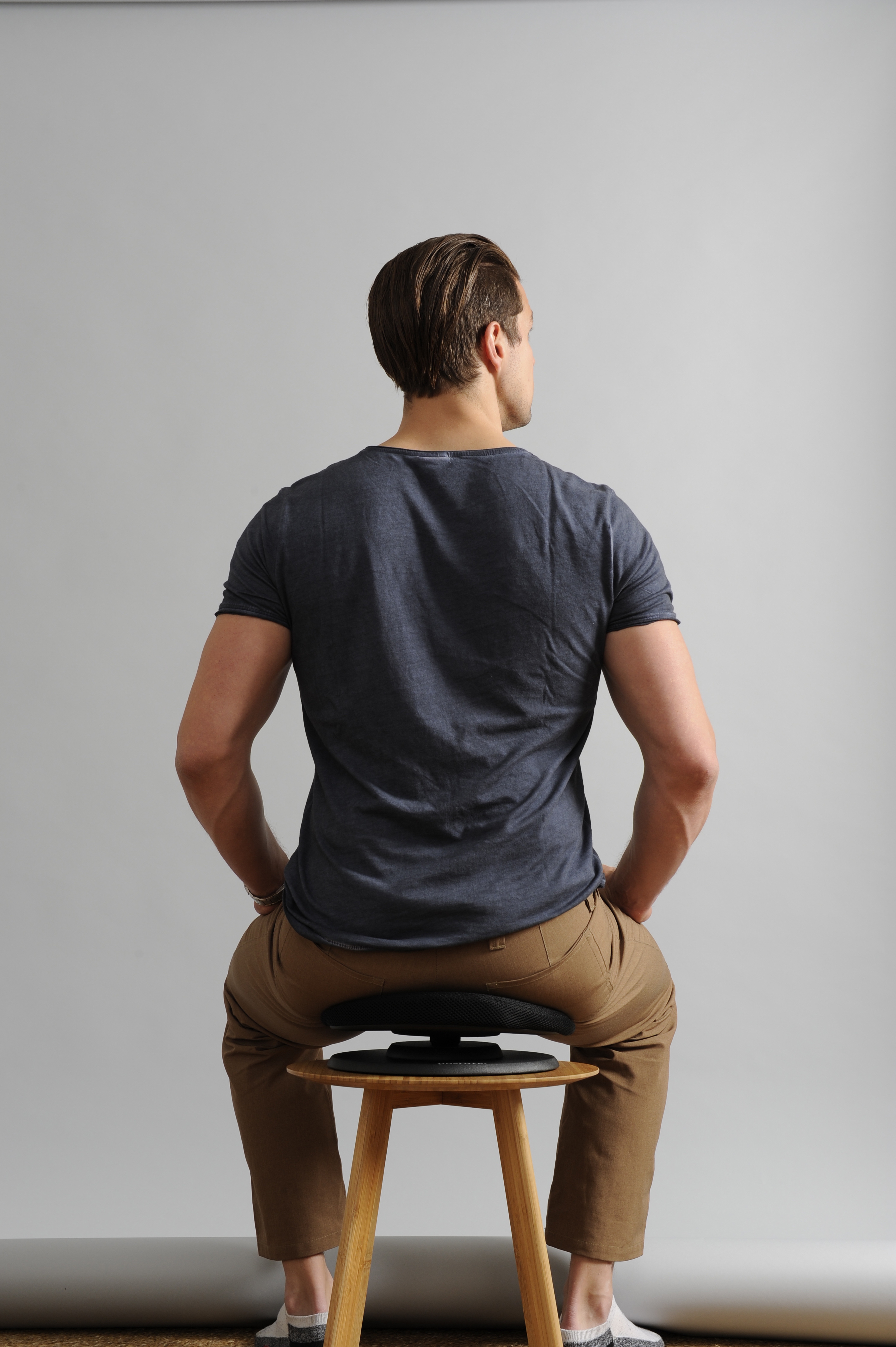 Ein Mann sitzt auf einem Hocker, dem Betrachter den Rücken zugewandt. Er trägt ein dunkles Hemd und braune Hose.