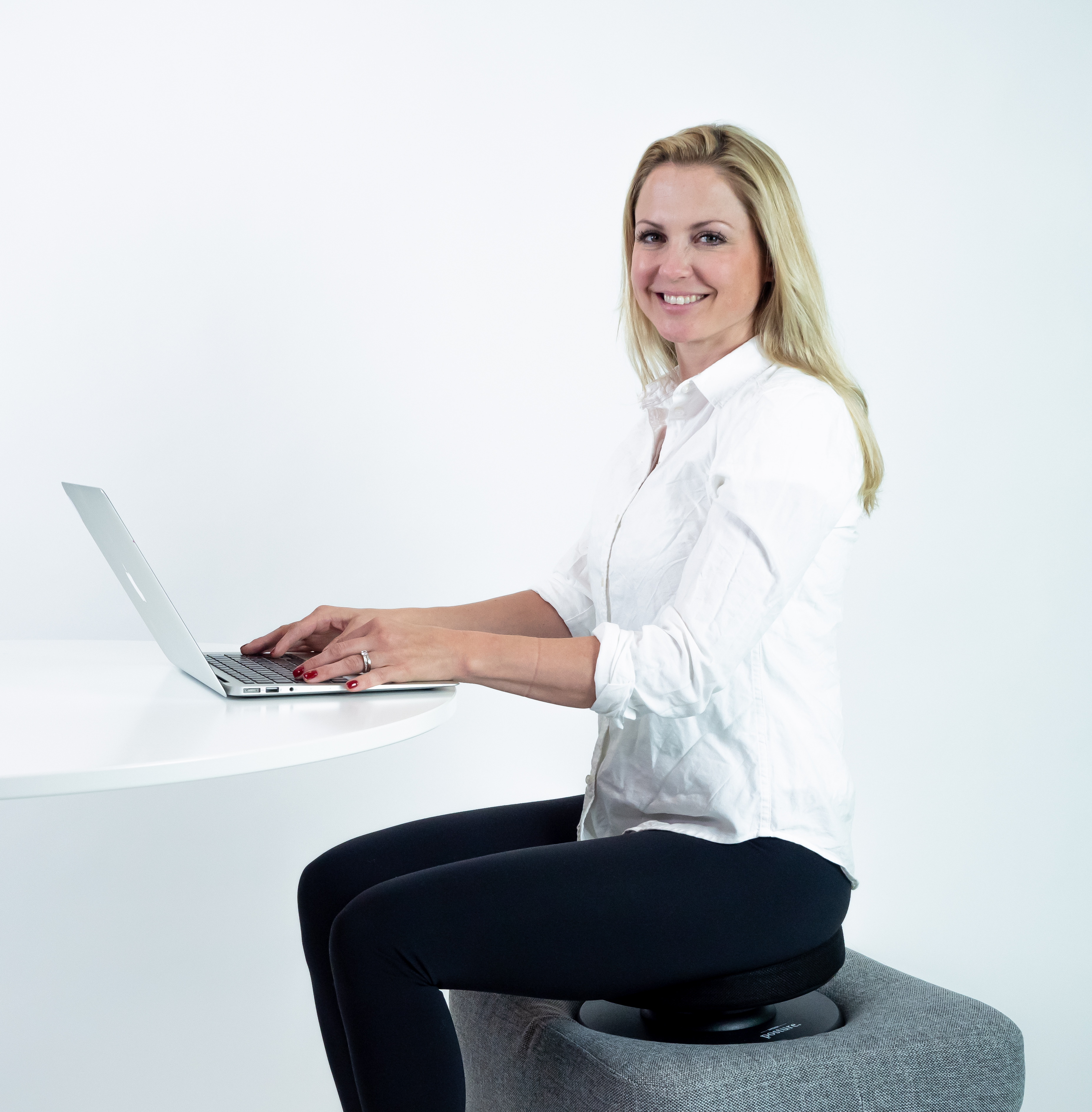 Frau sitzt an einem Tisch und benutzt einen Laptop. Sie lächelt. Weißer Tisch und Hintergrund.