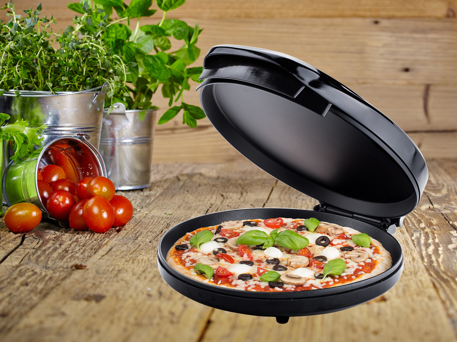Une pizza avec des tomates, des olives et du basilic dans un four à pizza noir, sur une table en bois.
