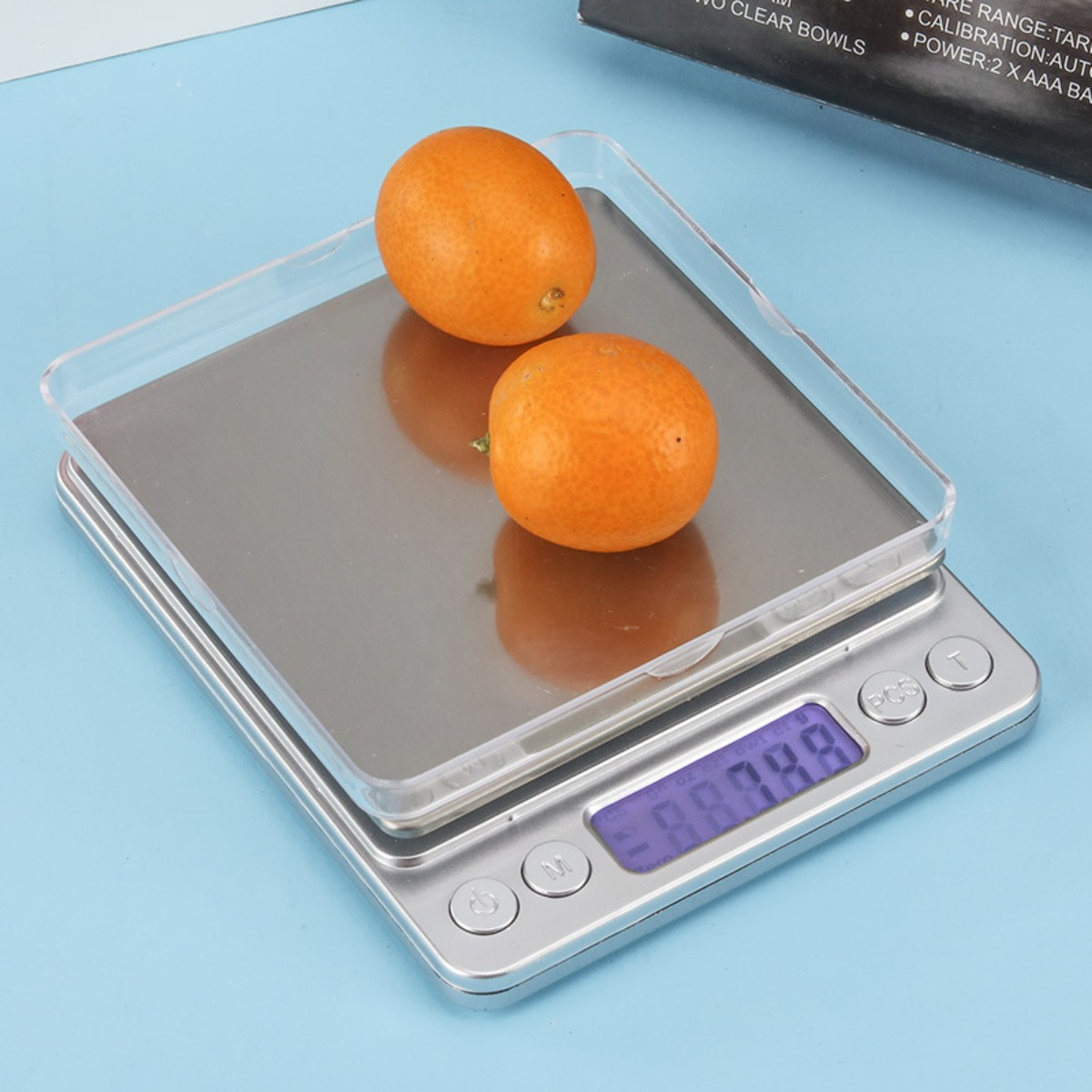 Silberne Digitalwaage mit zwei Orangen auf einem durchsichtigen Tablett; blauer Hintergrund.