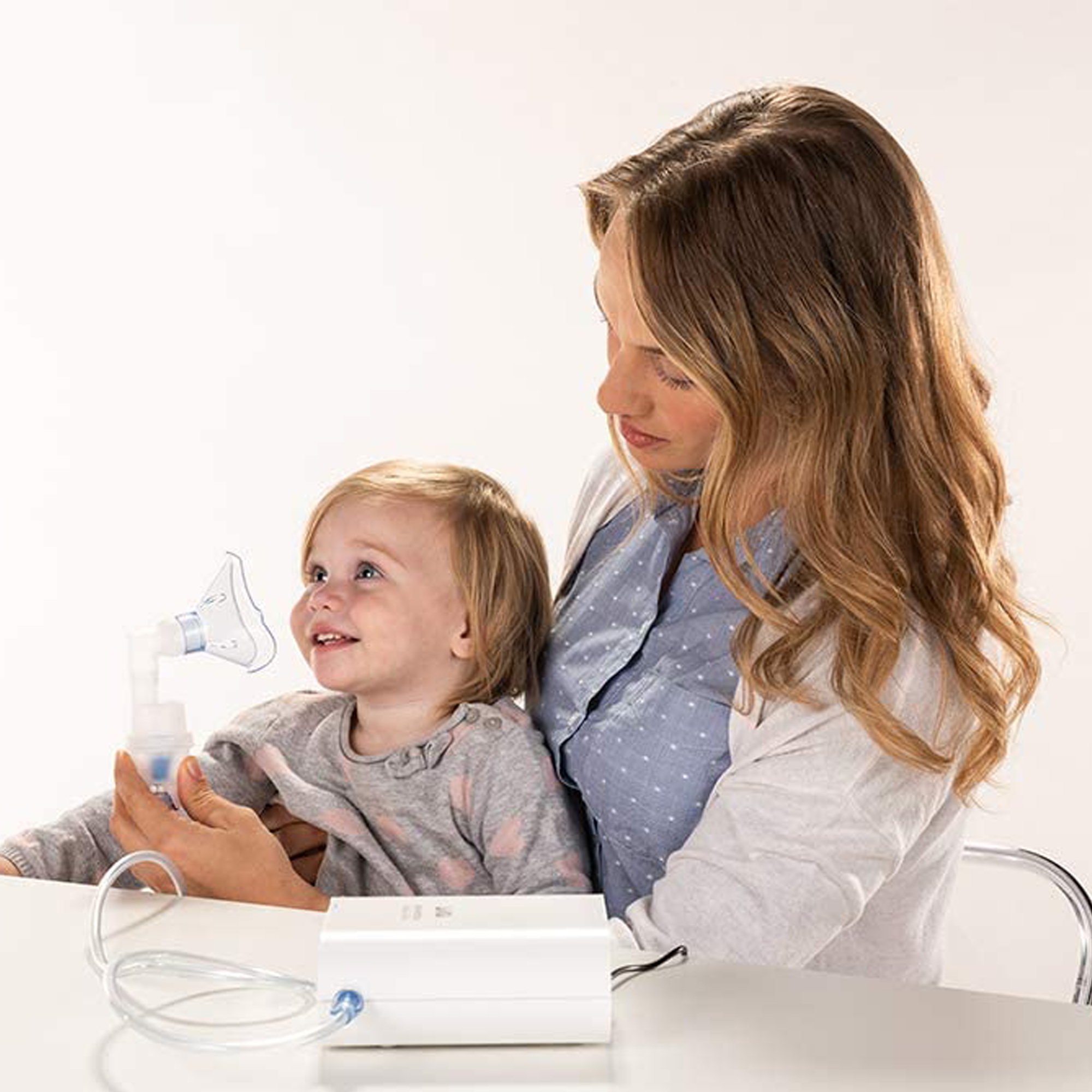 Una mujer sostiene a un bebé, que está usando un nebulizador. Están en una habitación blanca.