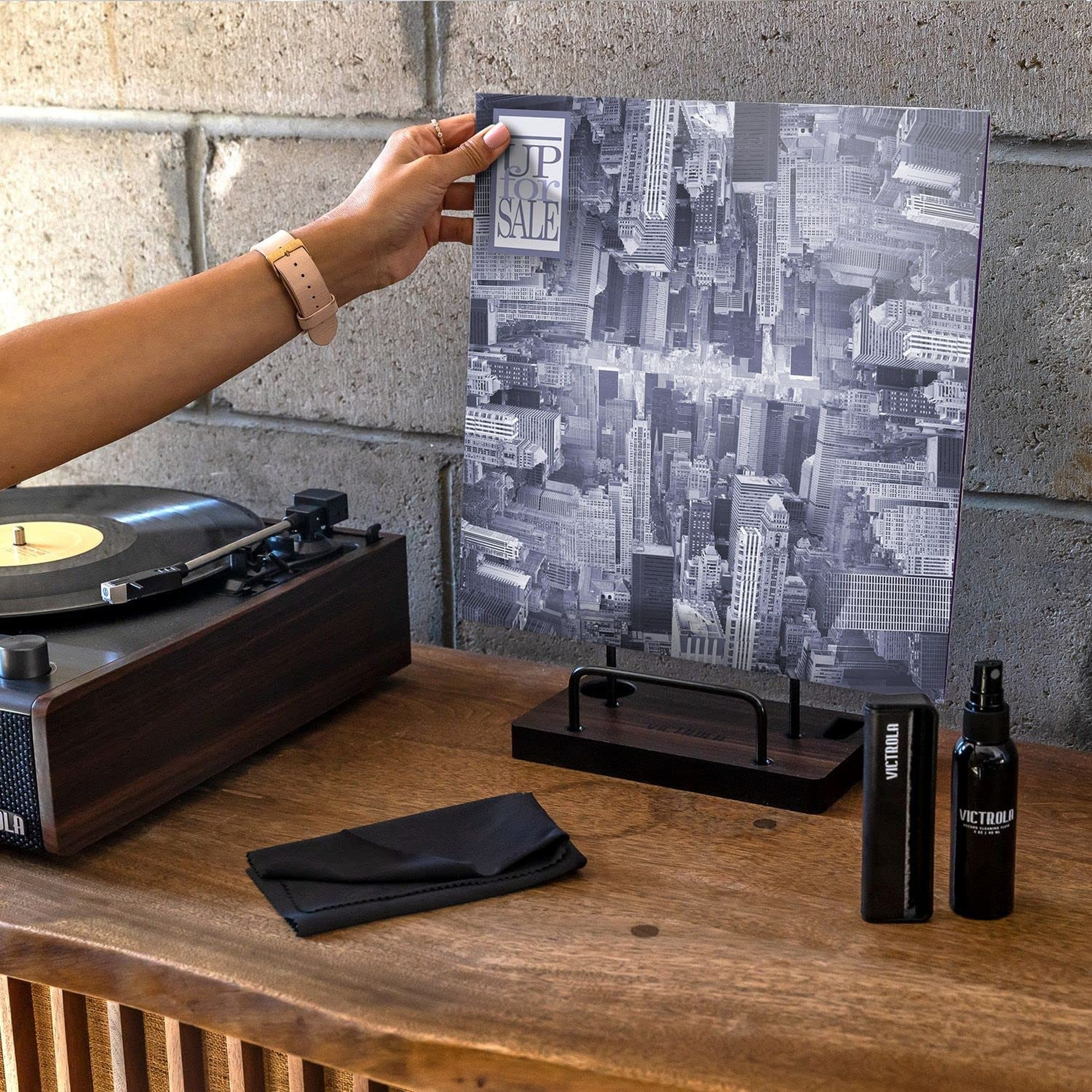 Persona sosteniendo una foto sobre un tocadiscos, con accesorios sobre una mesa de madera.
