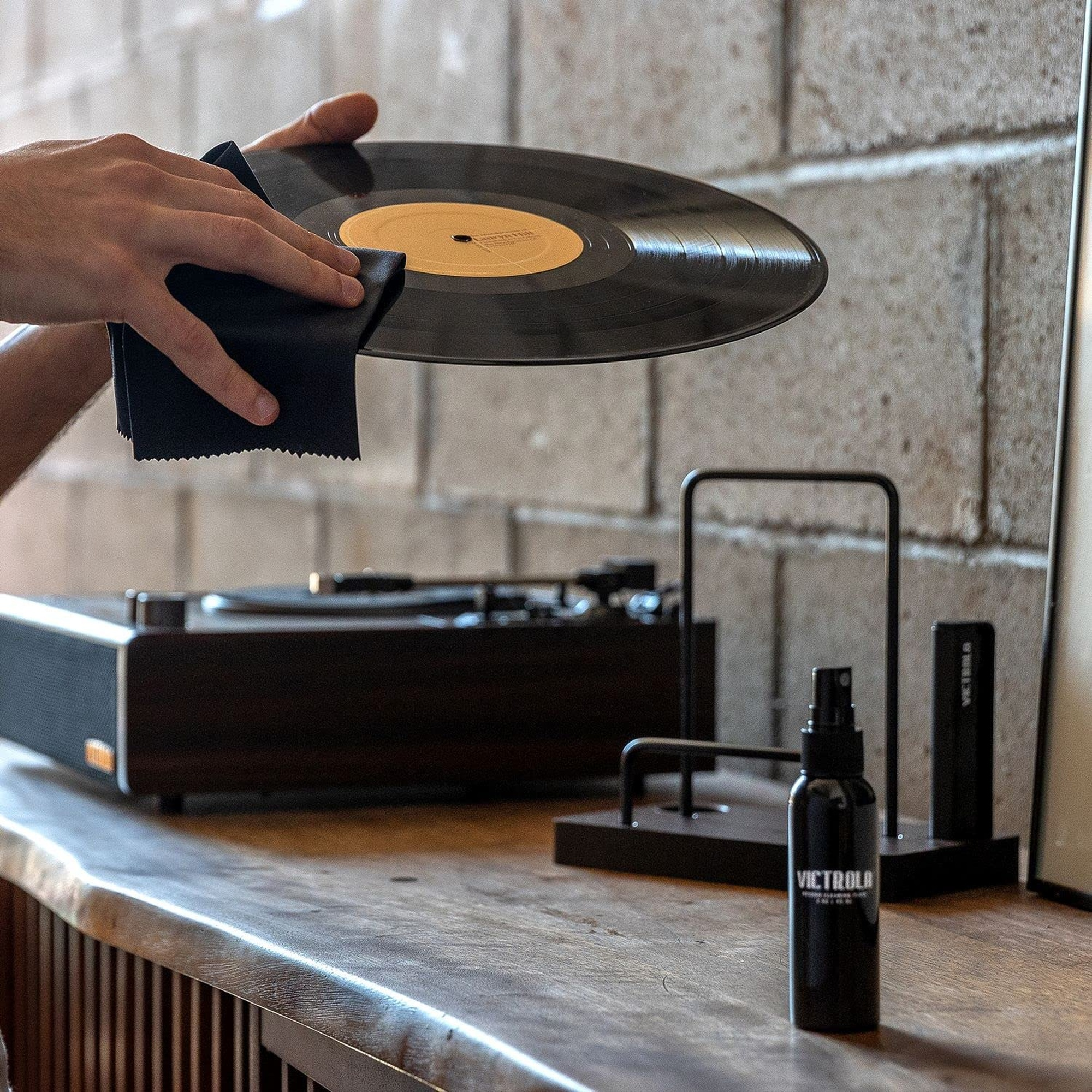 Persona limpiando un disco con un paño. Tocadiscos, altavoz, botella de spray y soporte para discos.