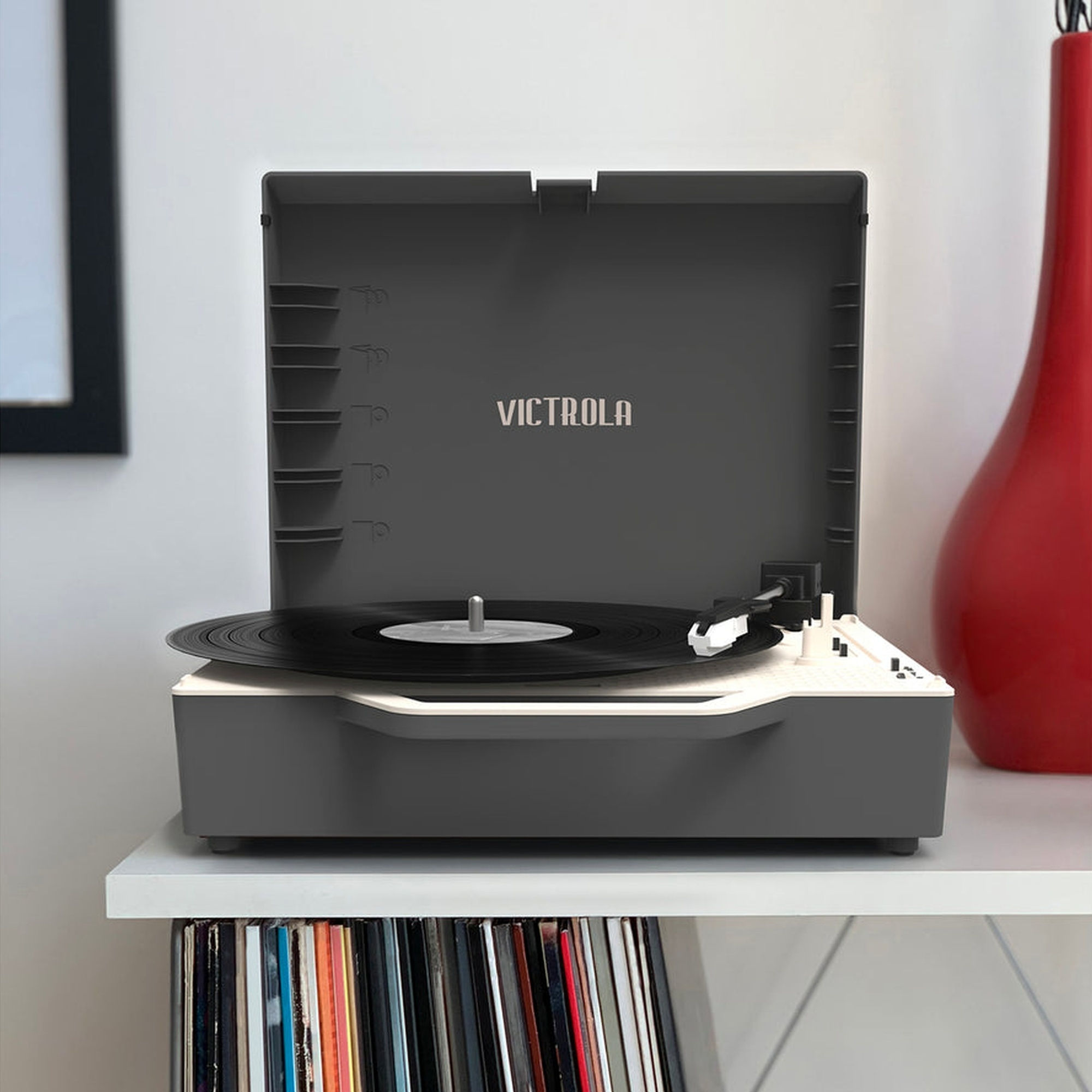 Un tocadiscos Victrola gris con un disco encima, sobre un estante blanco con discos debajo.