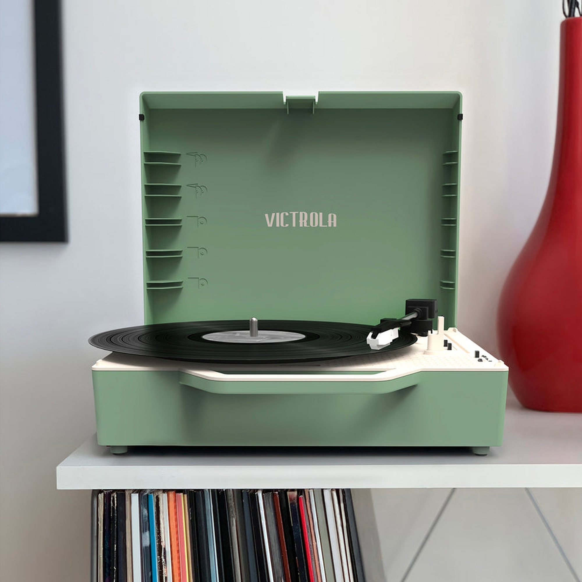 Tocadiscos Victrola verde abierto. Disco negro en el tocadiscos, estante blanco con discos.