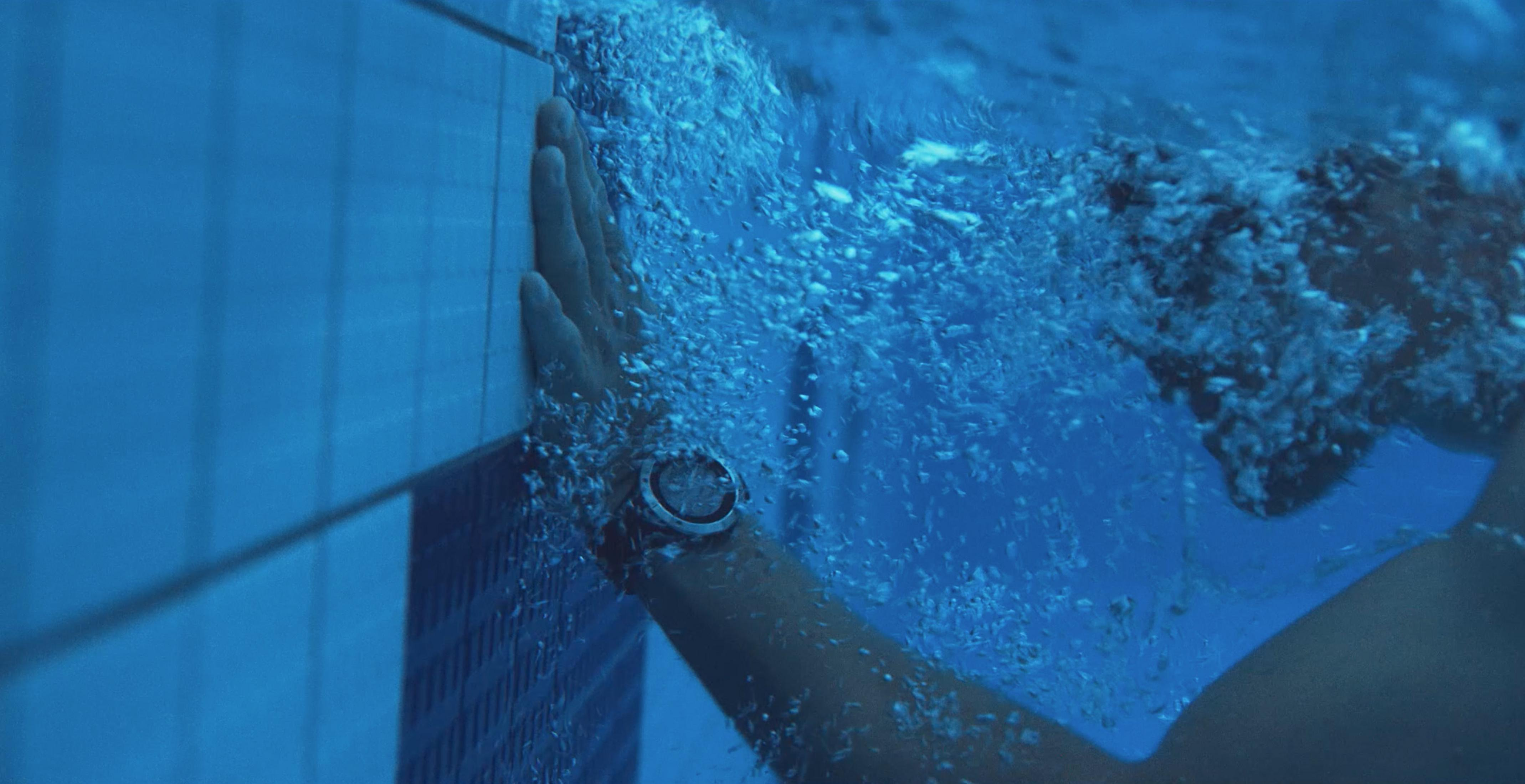 Hand einer Person mit Uhr in der Nähe einer blauen Fliesenwand unter Wasser mit Blasen.
