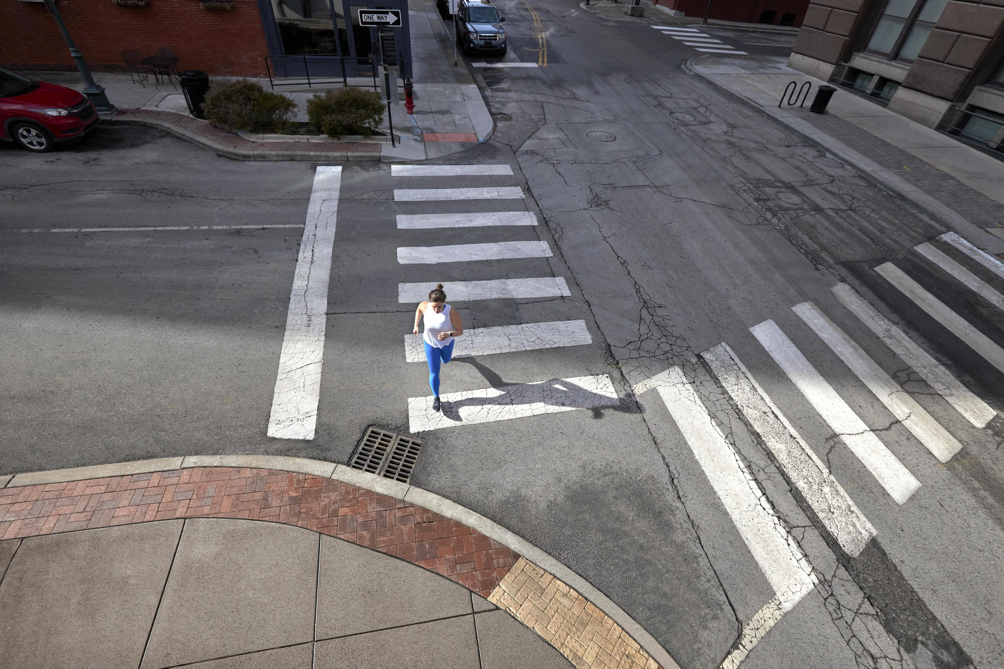 Biegacz w niebiesko-białym stroju przechodzi przez ulicę z przejściem dla pieszych. Samochody i budynki w tle.