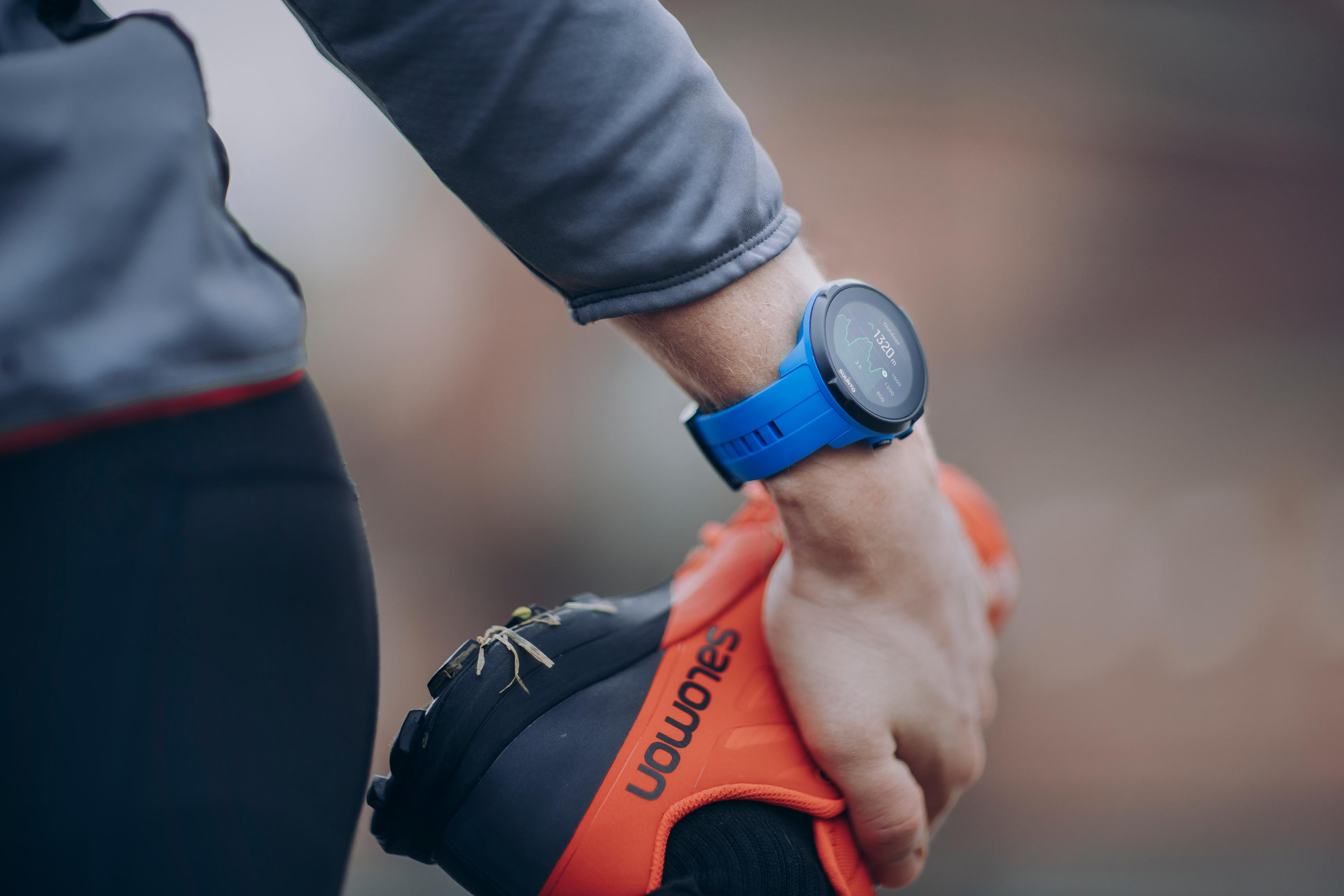 Eine Hand mit einer blauen Smartwatch, die einen orangefarbenen Laufschuh hält. Verschwommener Hintergrund.