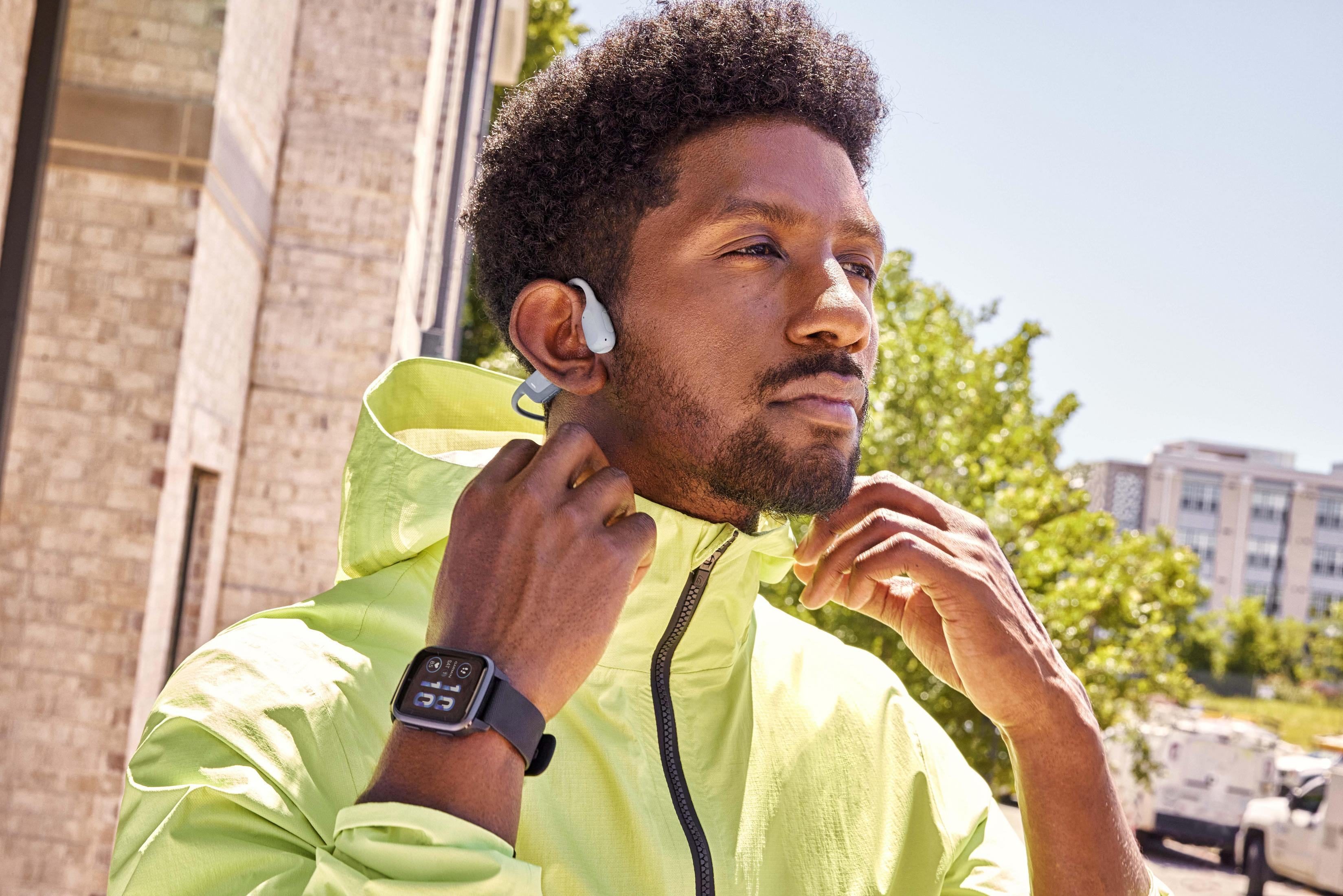 Mężczyzna w limonkowozielonej kurtce z słuchawkami i smartwatchem, na zewnątrz w słoneczny dzień.