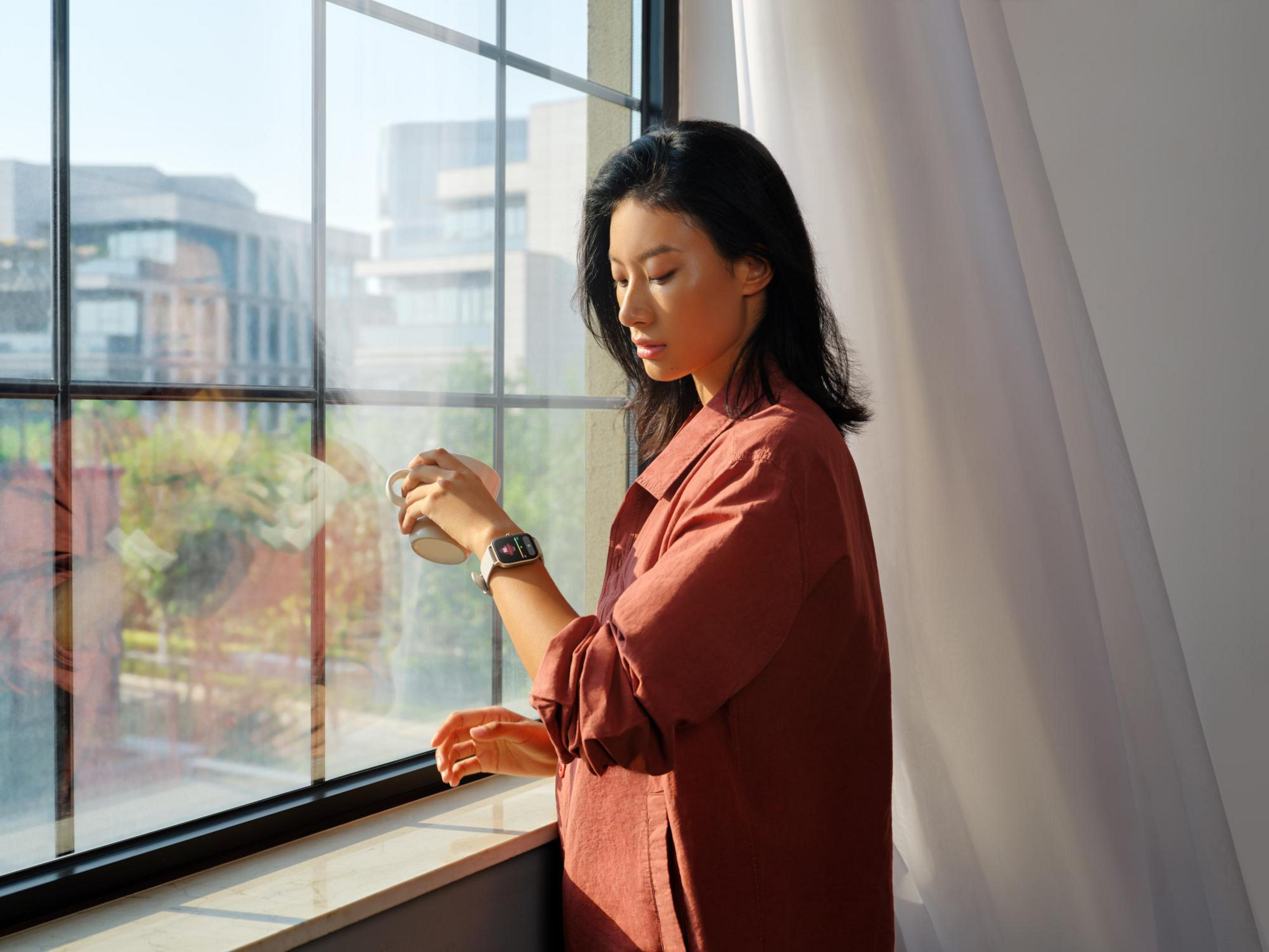 Mujer con camisa roja, reloj, mira por la ventana edificios.