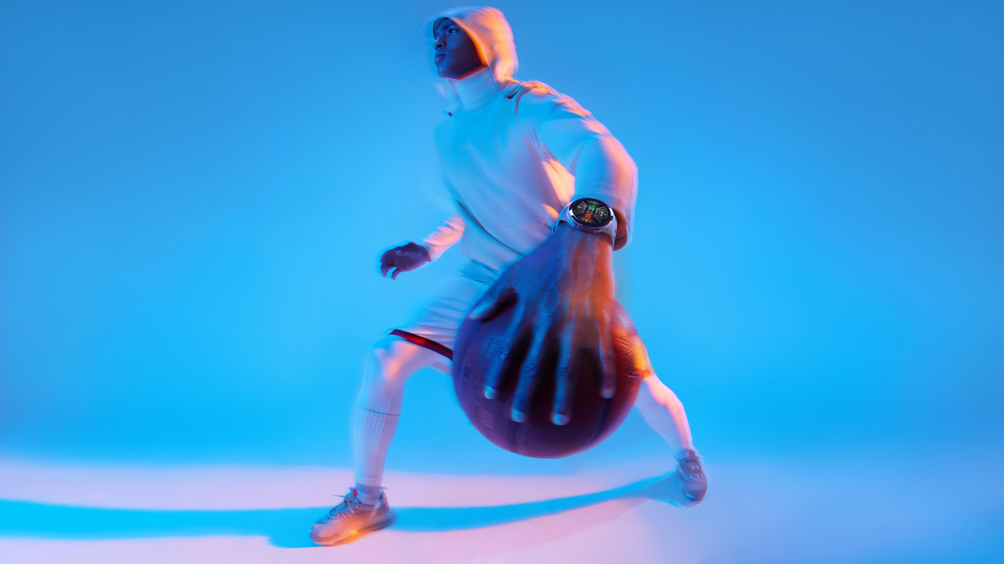 Sportowiec kozłuje piłkę do koszykówki, ubrany w białą bluzę z kapturem i zegarek na nadgarstku.