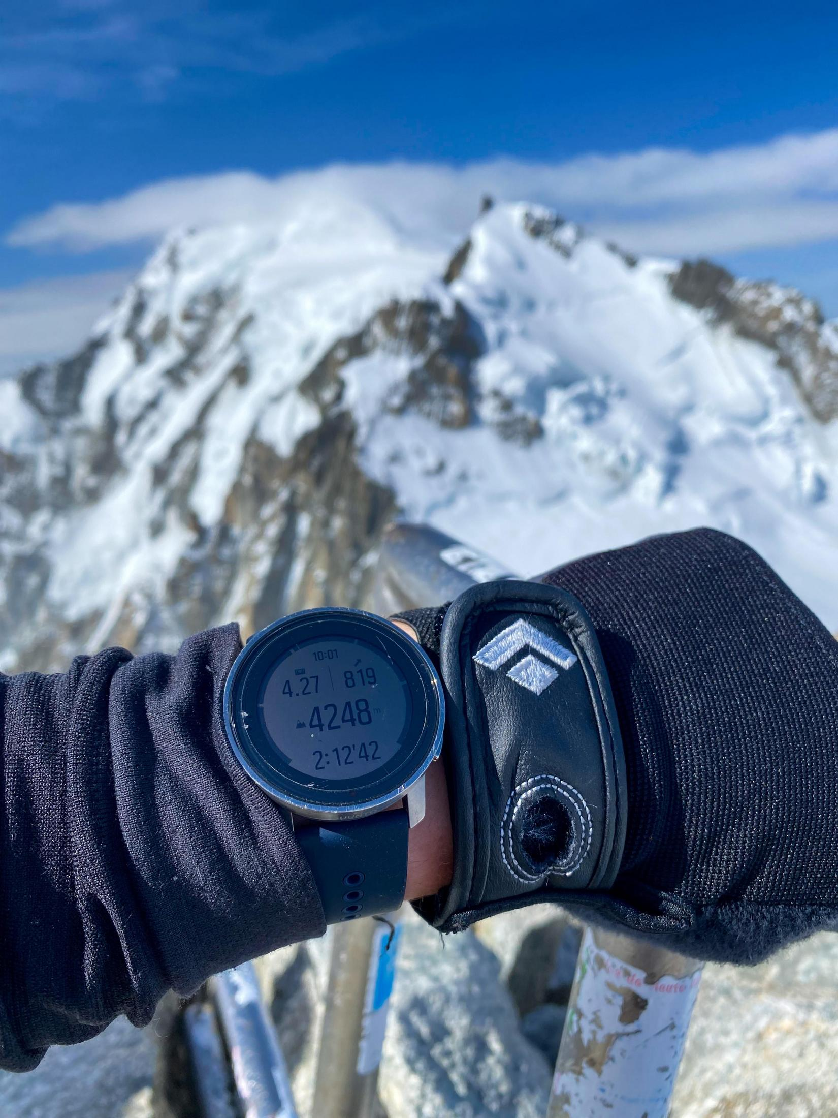 Muñeca con reloj inteligente mostrando altitud y hora, montaña nevada atrás.