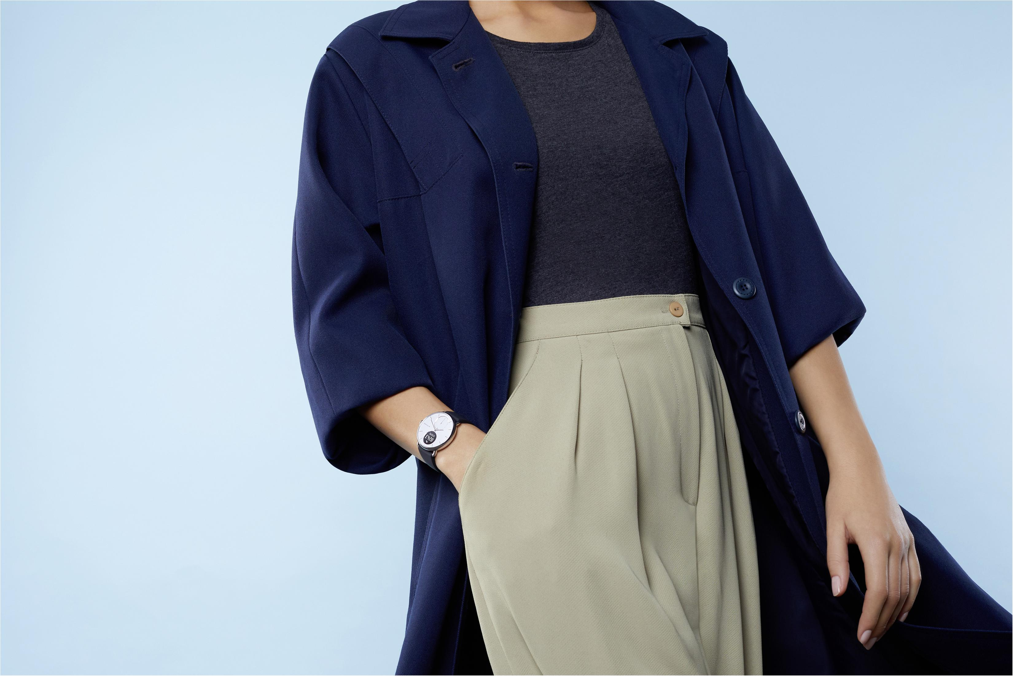 Eine Frau mit einem marineblauen Mantel, einem grauen Oberteil und einer beigen Hose, mit einer silbernen Uhr.