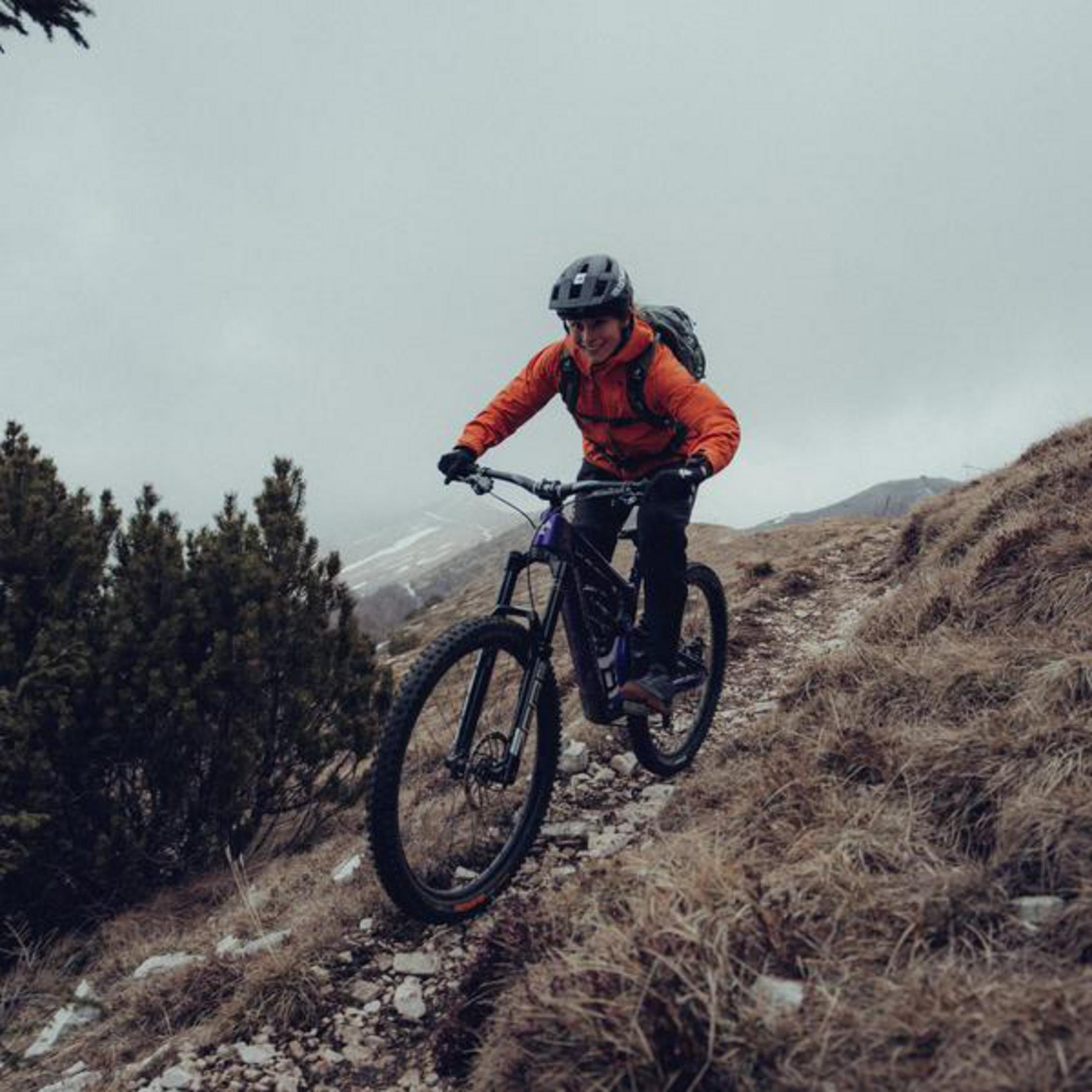 Osoba jadąca na rowerze górskim po polnej ścieżce, osoba ma na sobie kask i pomarańczową kurtkę.