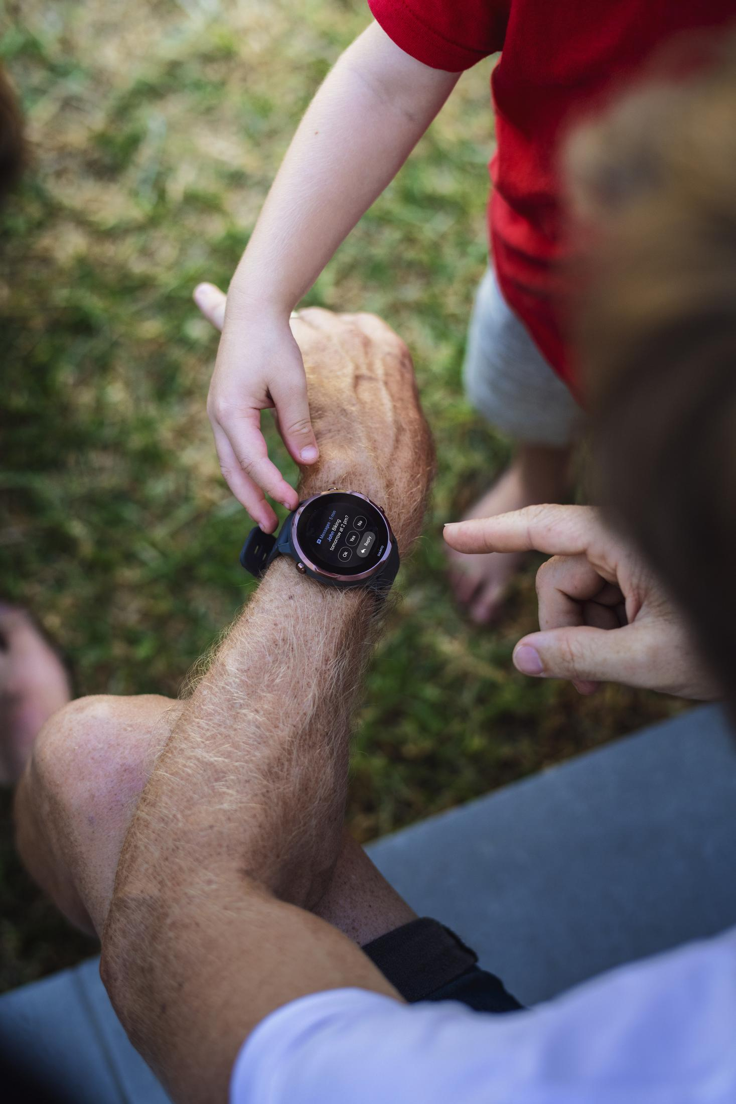 Eine Person trägt eine Smartwatch, ein Kind zeigt darauf. Szene ist im Freien.