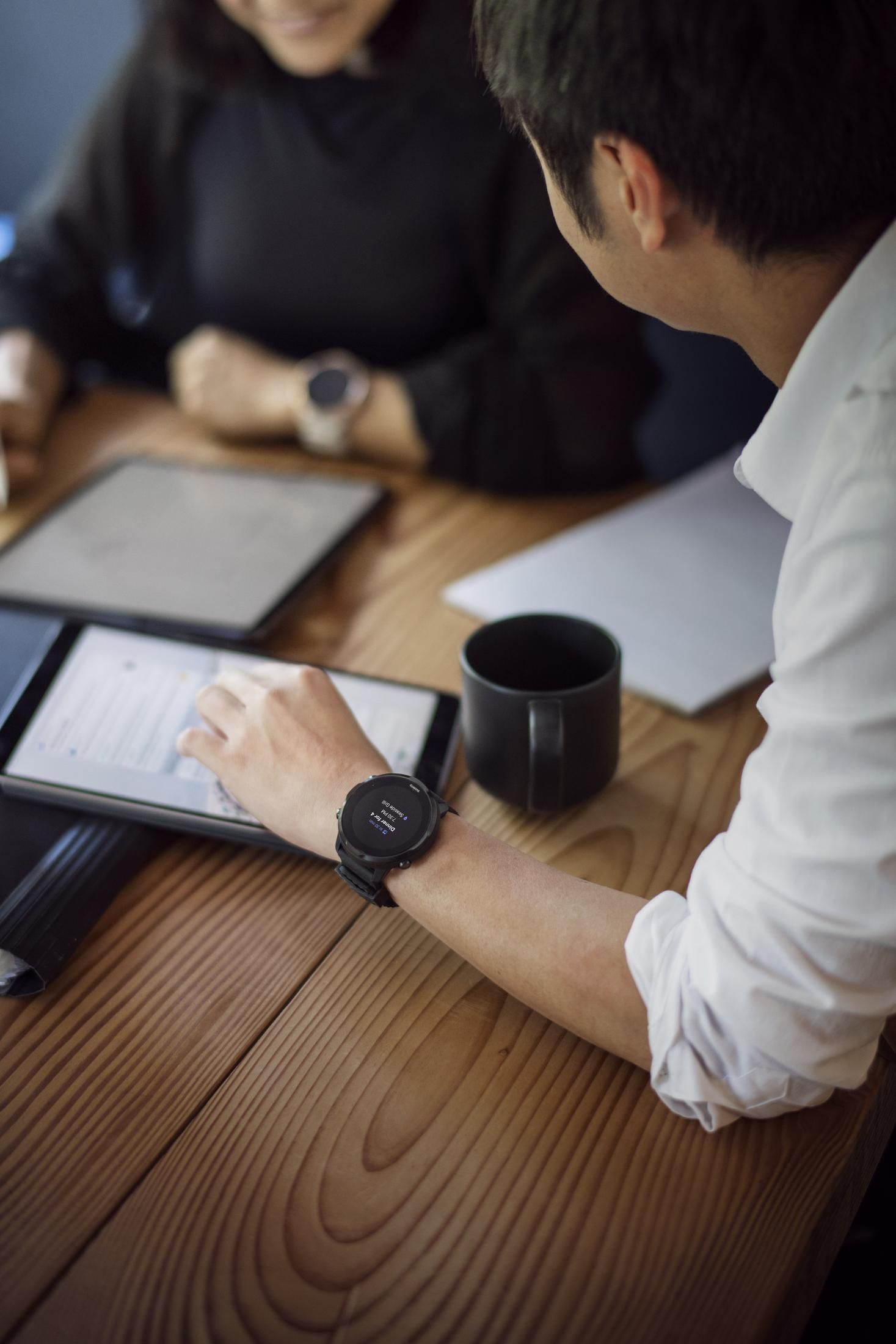 Zwei Personen betrachten Tablets, eine trägt eine schwarze Smartwatch, auf Holztisch.