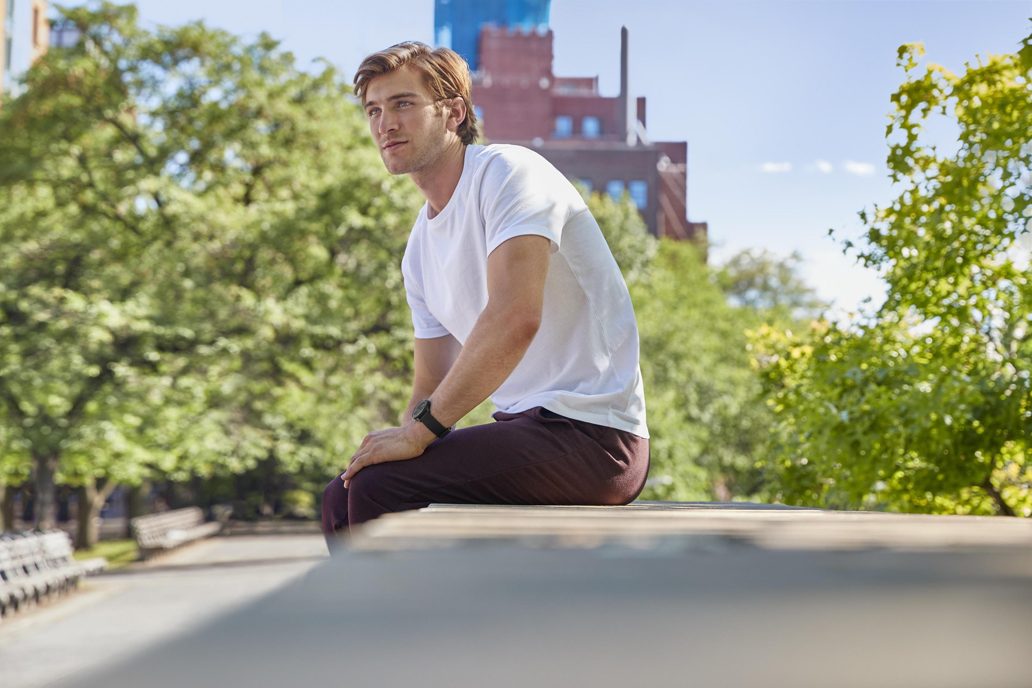 Mann sitzt auf einer Mauer in einem Park, blickt zur Seite. Bäume und Gebäude im Hintergrund.