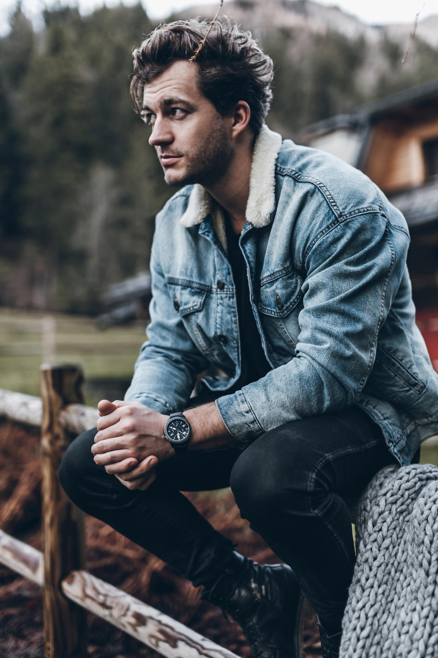 Mann in Jeansjacke sitzt auf Holzzaun, trägt Uhr, mit verschwommenem Hintergrund.