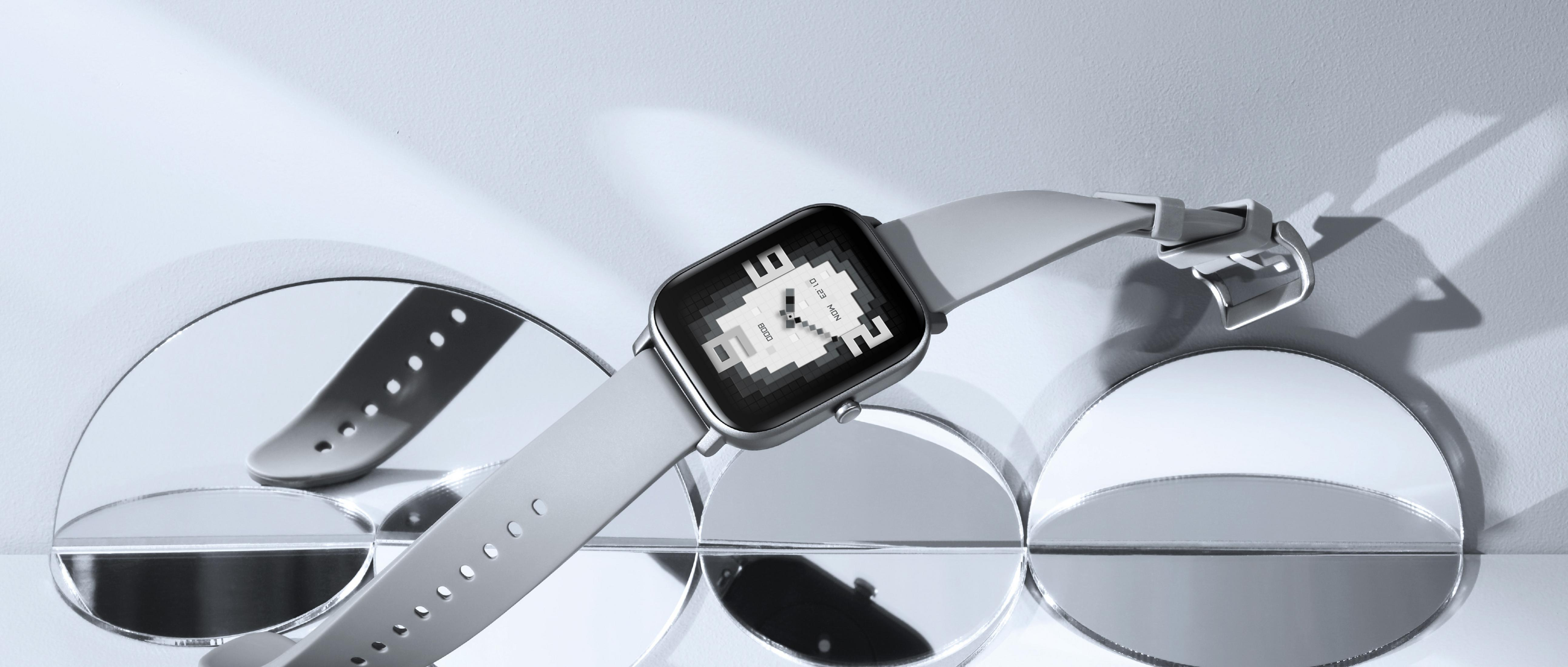 Uno smartwatch grigio chiaro con schermo quadrato e cinturino grigio chiaro su una superficie con specchi.