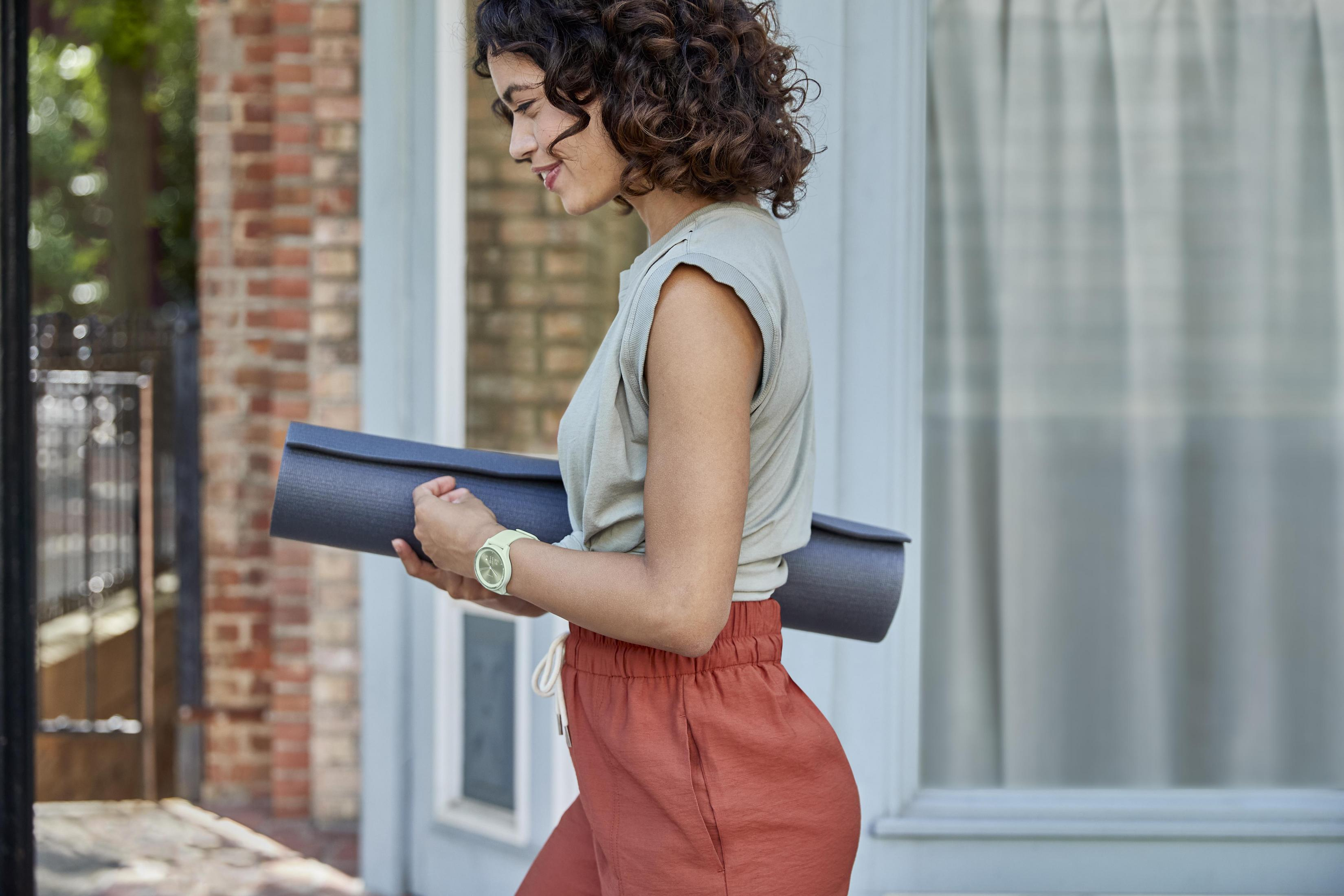 Frau mit lockigem Haar, hält eine Yogamatte und geht neben einem Backsteingebäude entlang.