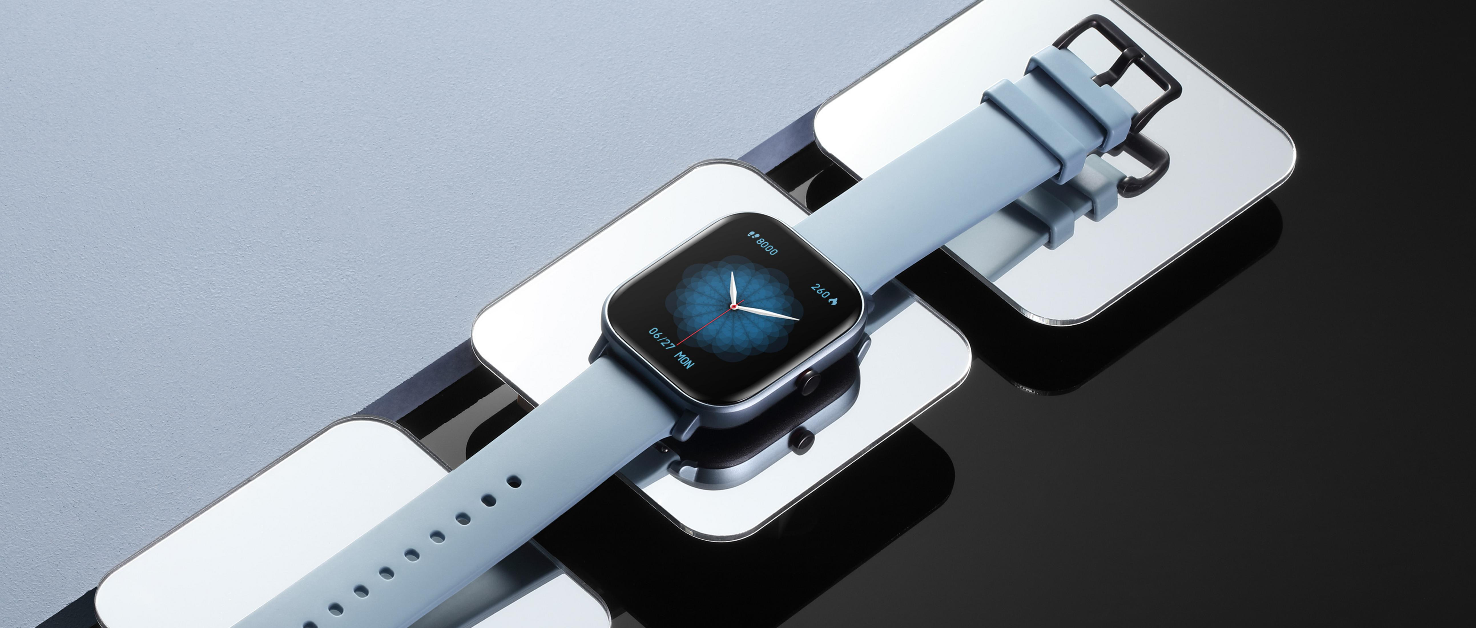 Uno smartwatch azzurro con un quadrante quadrato, appoggiato su superfici riflettenti.