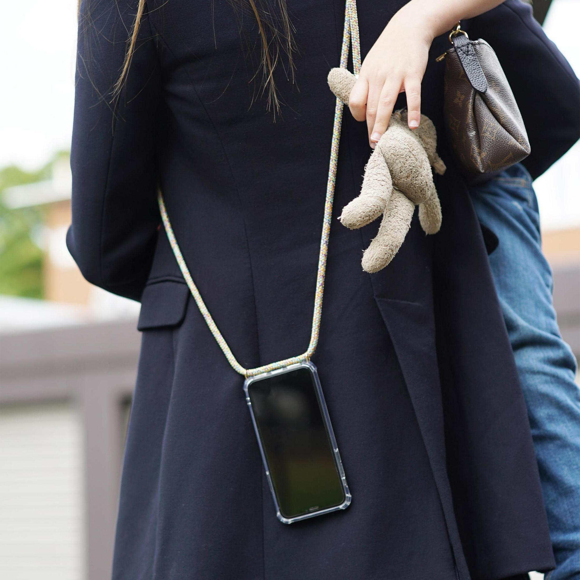 Person trägt schwarze Jacke, hält Telefon an einem Lanyard und Stofftier, mit Kind.