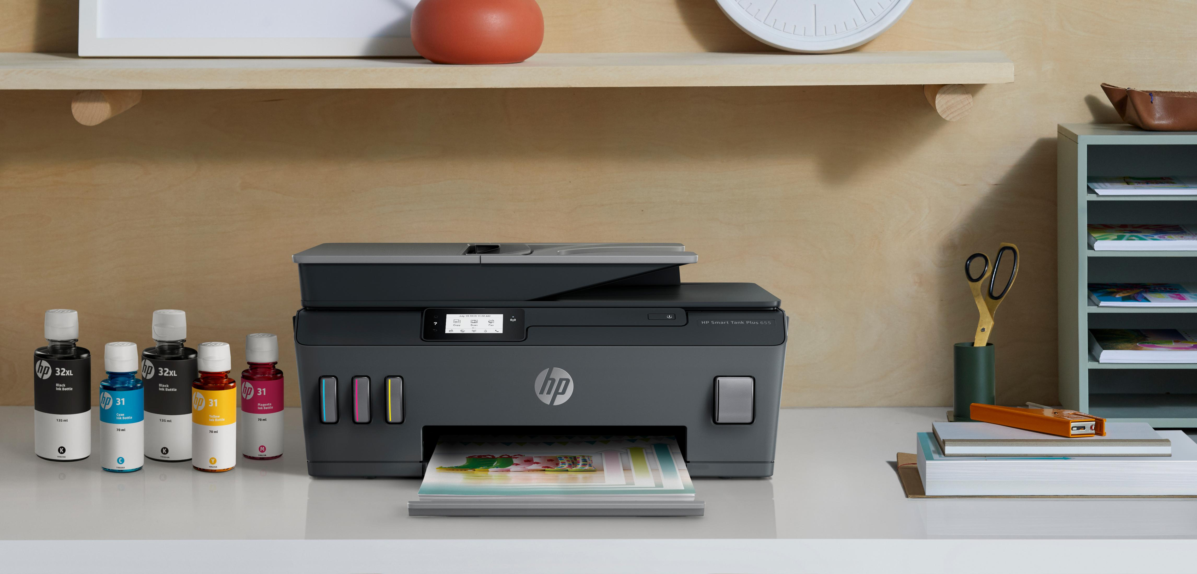 Drukarka HP z butelkami z tuszem i materiałami eksploatacyjnymi. Na biurku z drewnianą półką w tle.