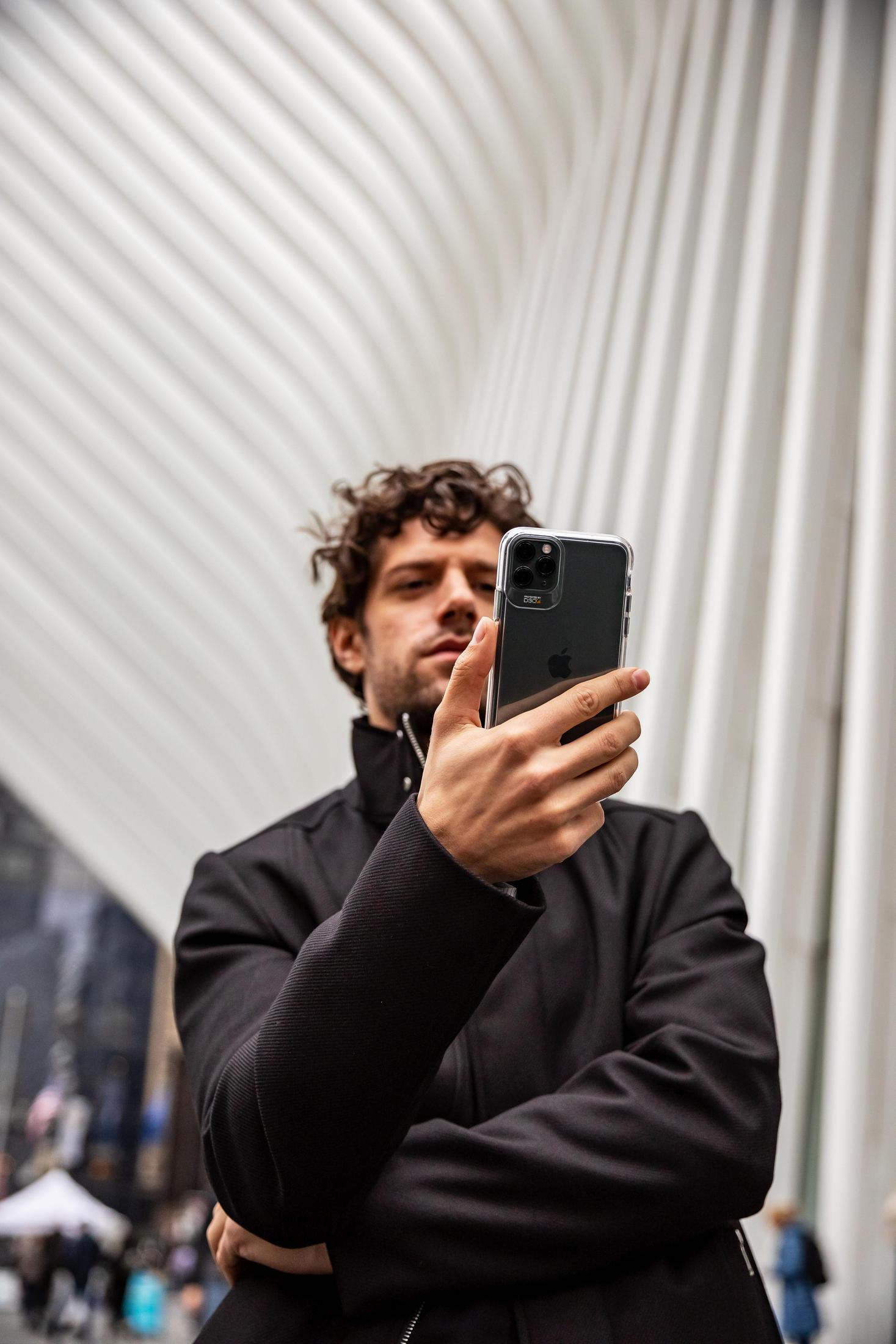Un uomo che tiene un telefono davanti a un edificio moderno, sfondo sfocato.