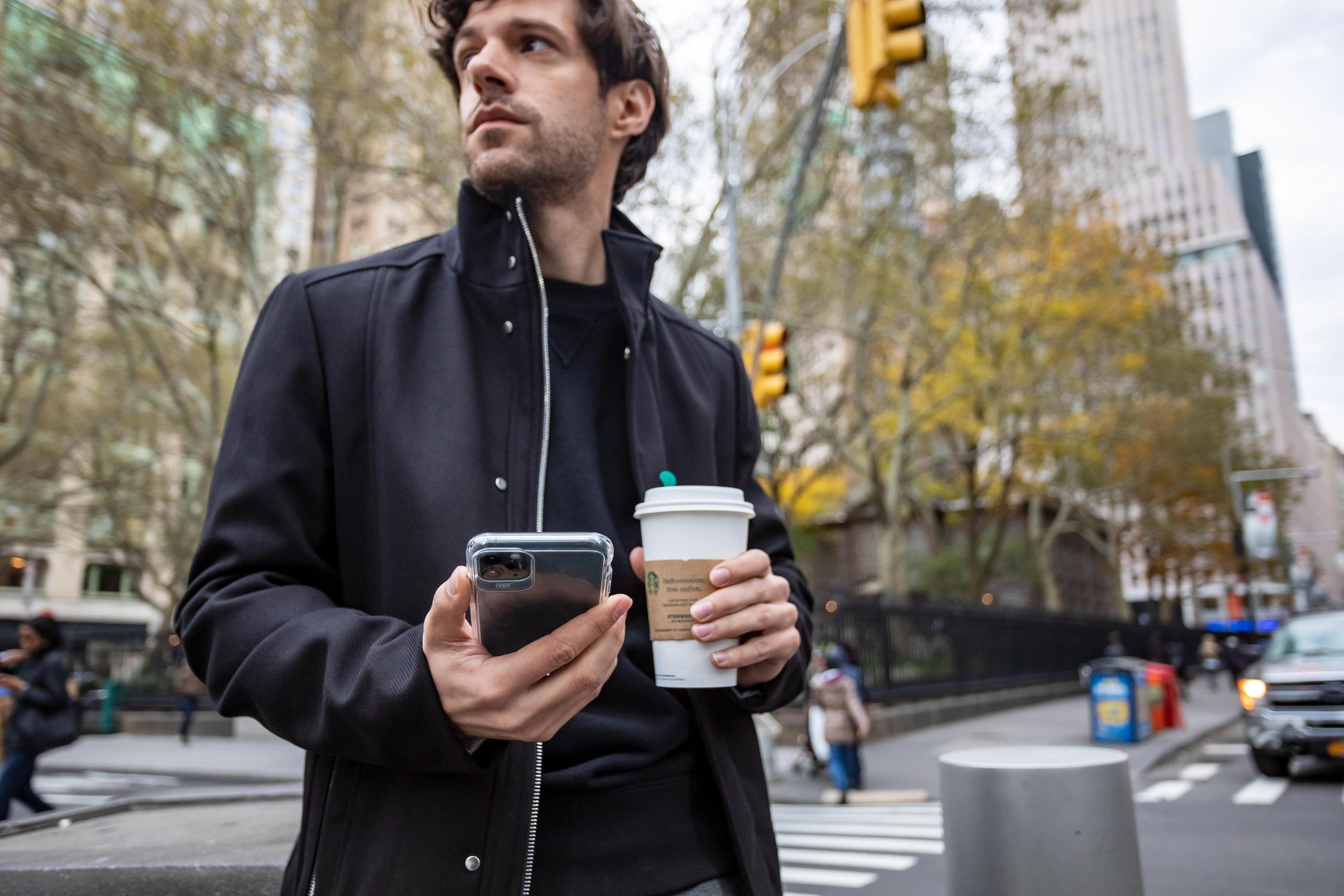 Uomo in giacca nera, con caffè e telefono, in piedi in una città.