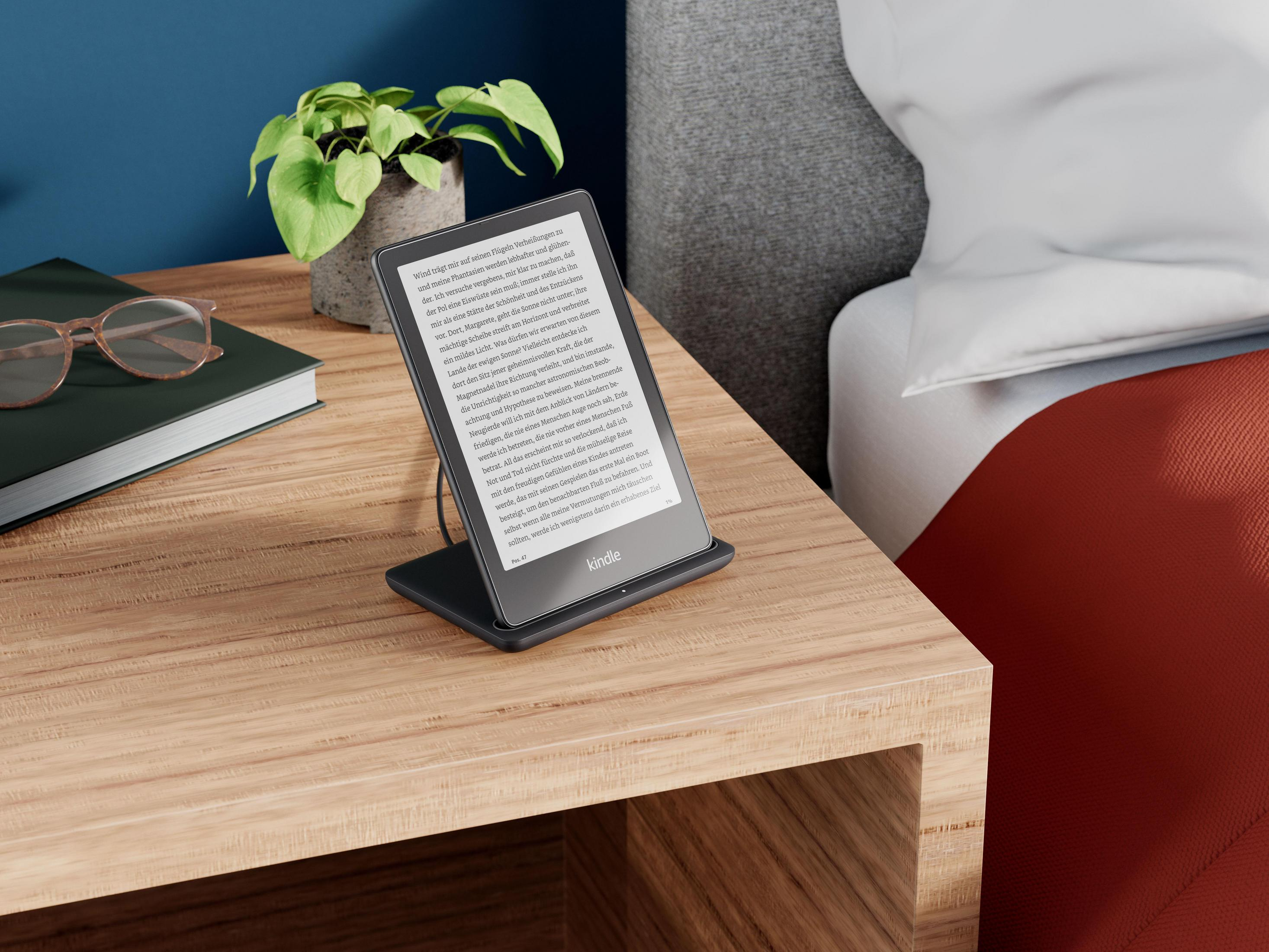 Ein Kindle-E-Reader auf einem Ständer, auf einem hölzernen Nachttisch, mit einer Pflanze und einem Buch.